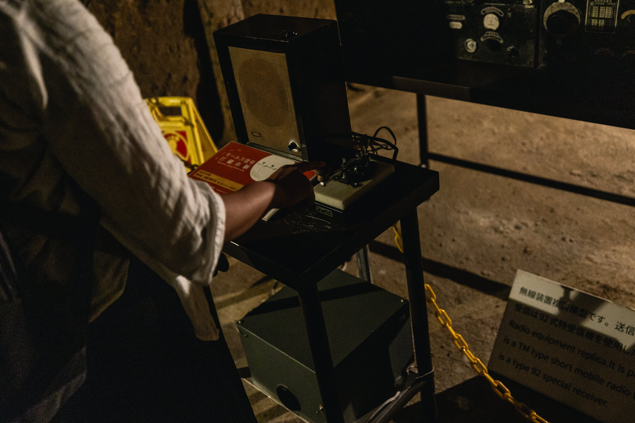 A visitor’s hand on a telegraph key beside a speaker and a red Morse code booklet, with a sign at the lower right reading “Radio equipment replica”