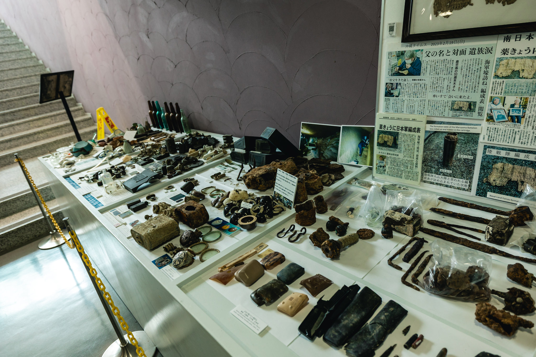 A long display table of recovered belongings — bottles, combs, lenses and rusted metal — with newspaper clippings and photographs of a rolled document on the wall at the right