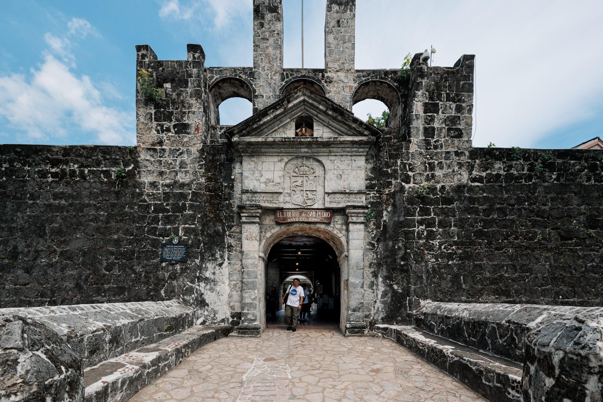 The stone front gate of Fort San Pedro, with a wooden sign reading EL FUERTE de SAN PEDRO 1565 over the arched entrance, a carved coat of arms and inscriptions above it, and a historical plaque on the wall to the left