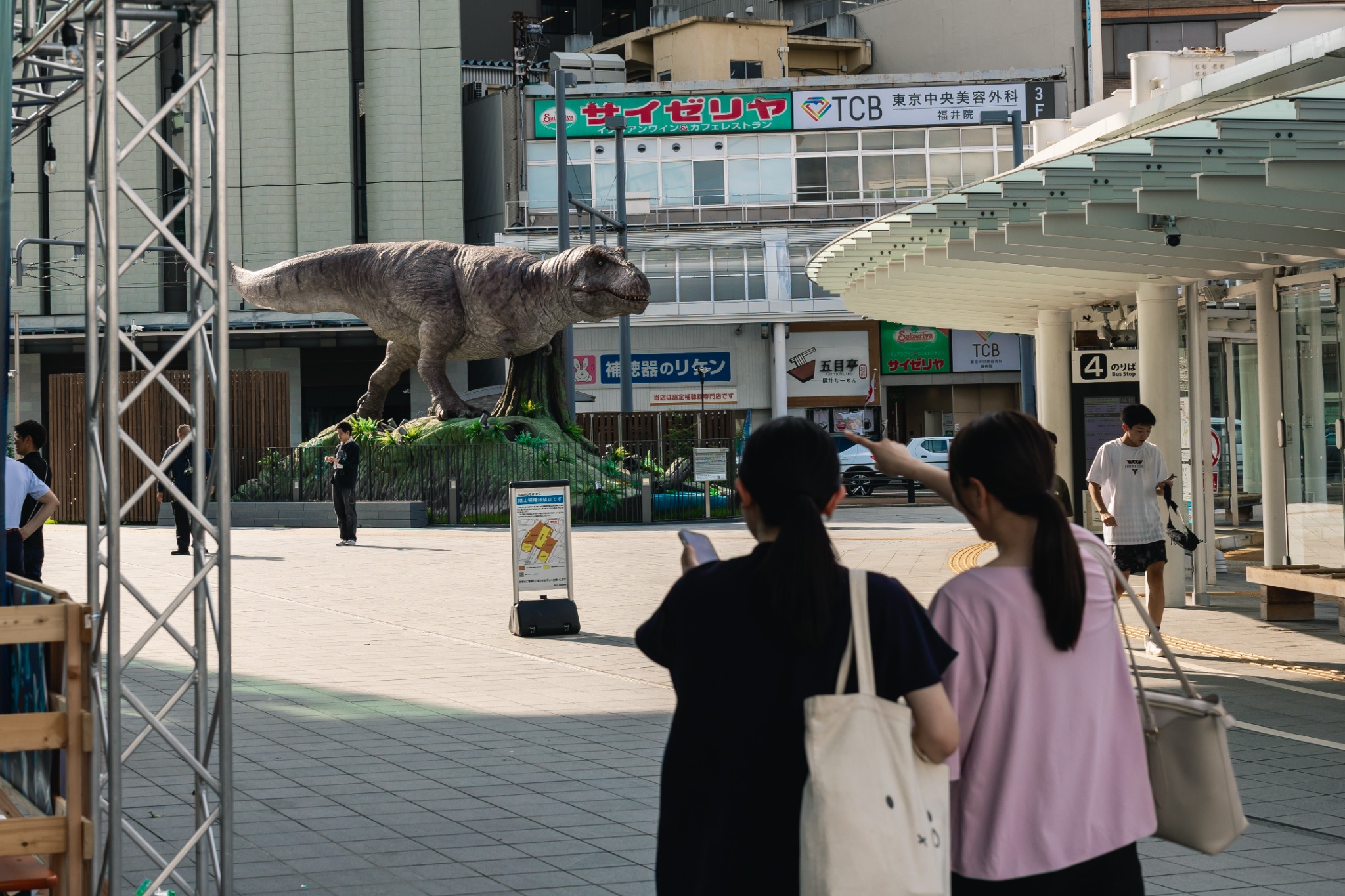 Large animatronic Tyrannosaurus outside Fukui Station, with two visitors in the foreground, one pointing toward it.