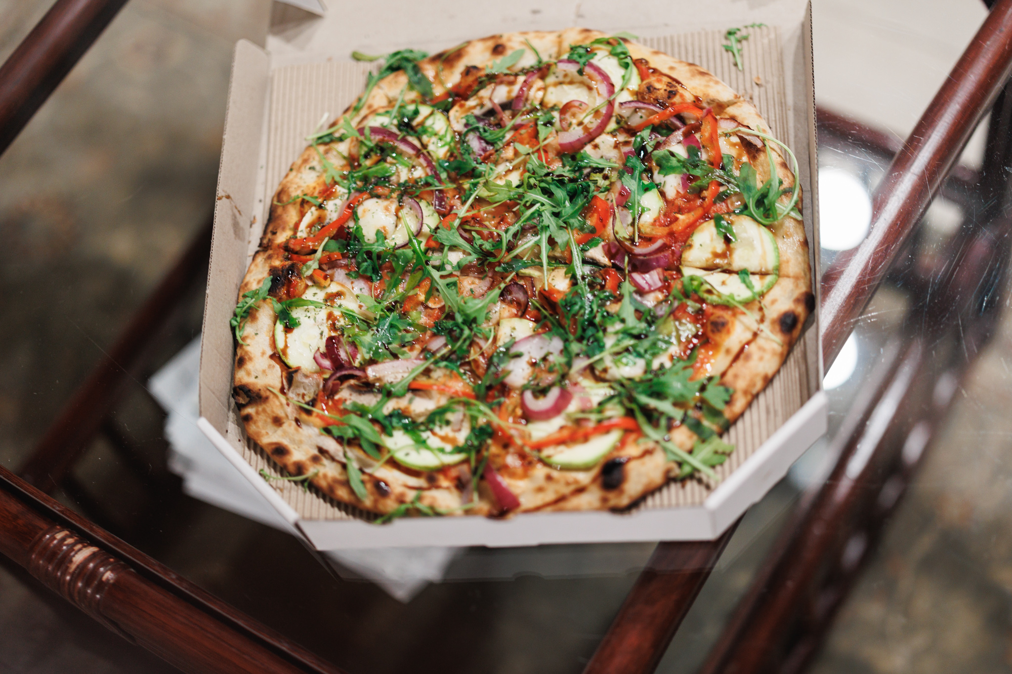 A round vegetable pizza in an open cardboard box on a glass-topped table, covered with courgette slices, red onion, red pepper and rocket, its rim spotted with char