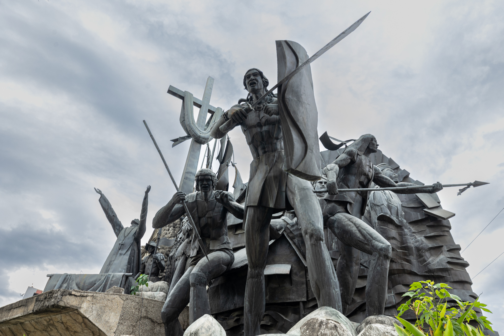 Low-angle view of dark sculpted warriors: one in the centre, mouth open, raising a sword beside a tall shield, another crouching with a spear, and a robed figure with both arms raised in front of a tall cross