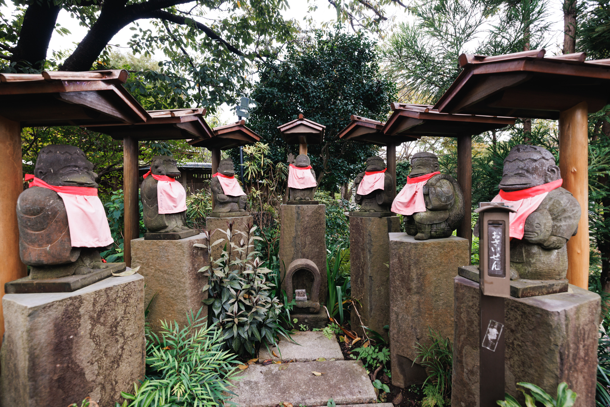 A row of stone kappa figures in pink aprons on stone pedestals under small wooden roofs, lining both sides of a garden path