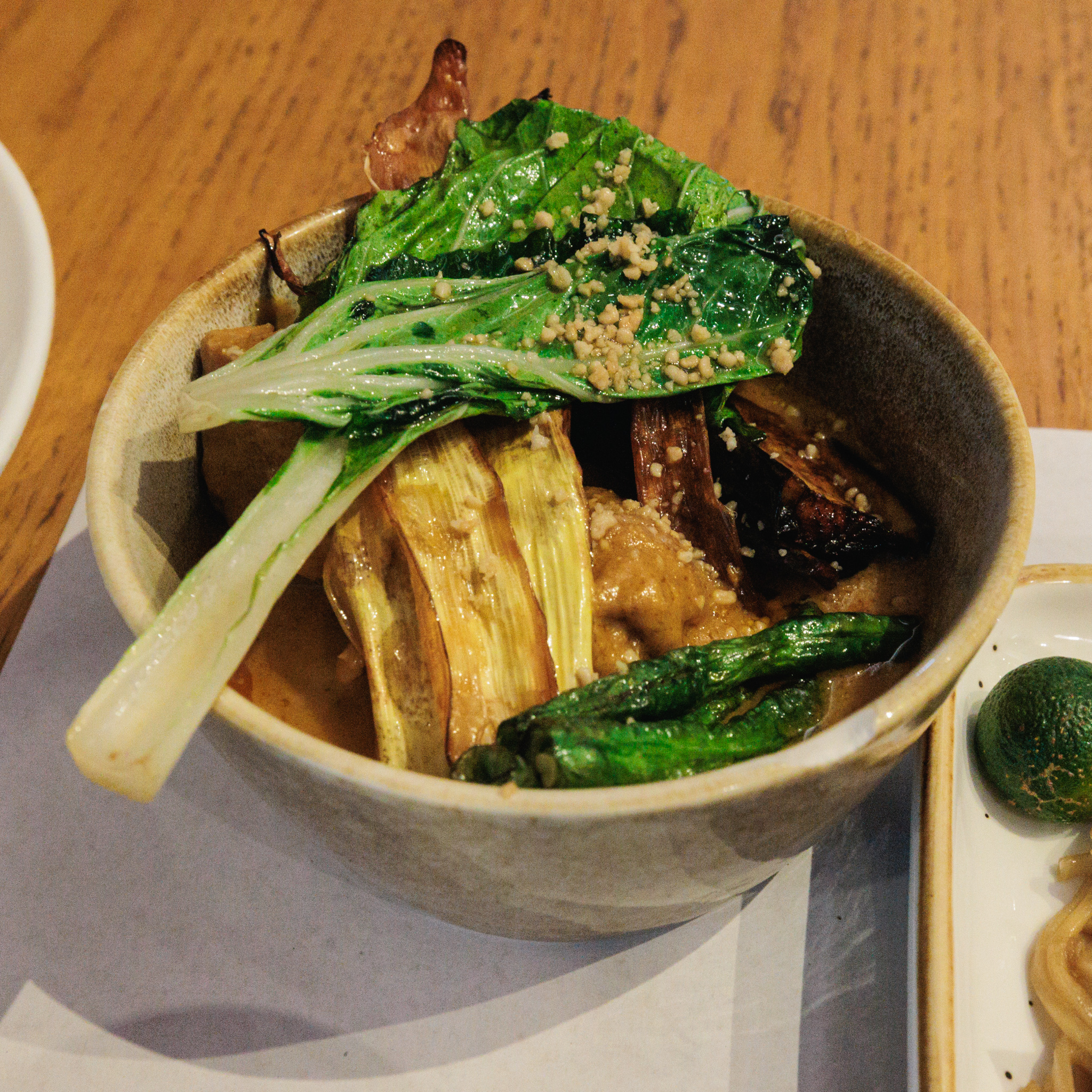 Close-up of kare-kare in a stoneware bowl: a glossy pechay leaf sprinkled with crushed peanuts lies over strips of eggplant, green beans and pieces of meat in a thick peanut sauce.