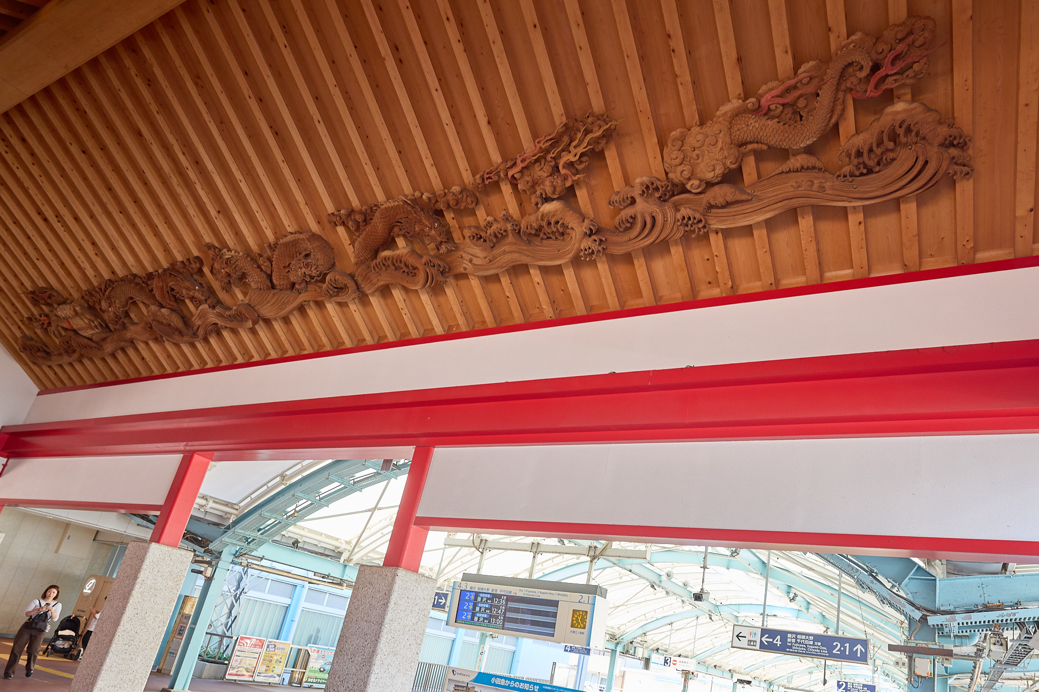 A long carved wooden dragon among clouds and waves on a slatted wooden ceiling above a red beam, with a departure board and platform signs beyond the opening below
