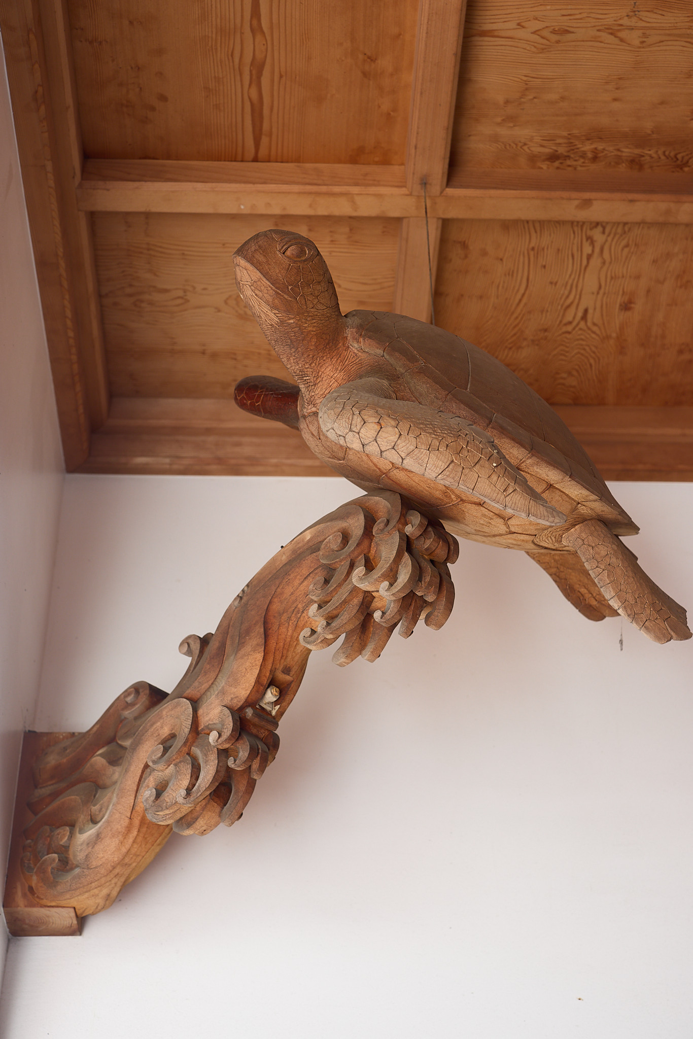 A carved wooden sea turtle with a patterned shell and outstretched flippers, riding on a carved wave bracket just below a wooden ceiling