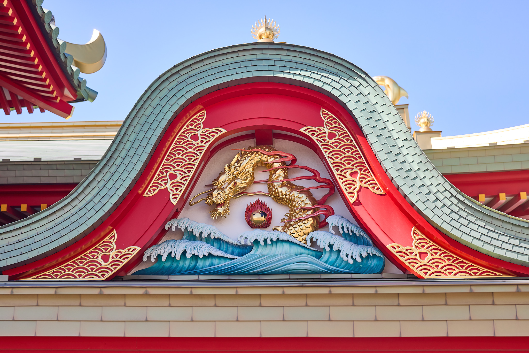 A gilded dragon over blue and white waves beside a red flaming ball, set in a red curved gable under green tiles and gold trim