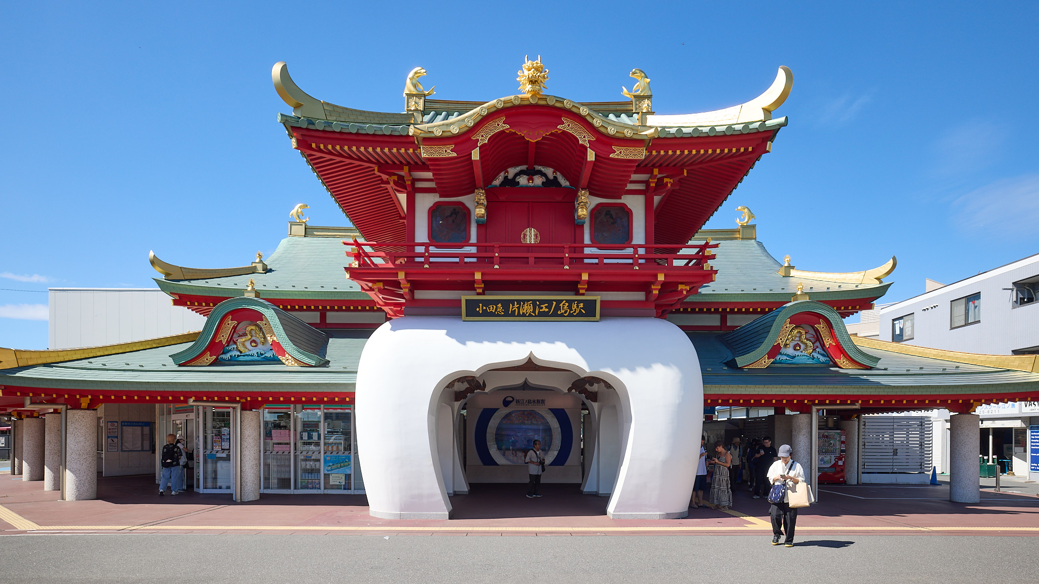 The front of Katase-Enoshima Station: a red palace-style gate tower with green and gold roofs and gilded dragon heads, standing on a white curved archway, with the station name board above the arch and a gilded dragon relief in each side wall