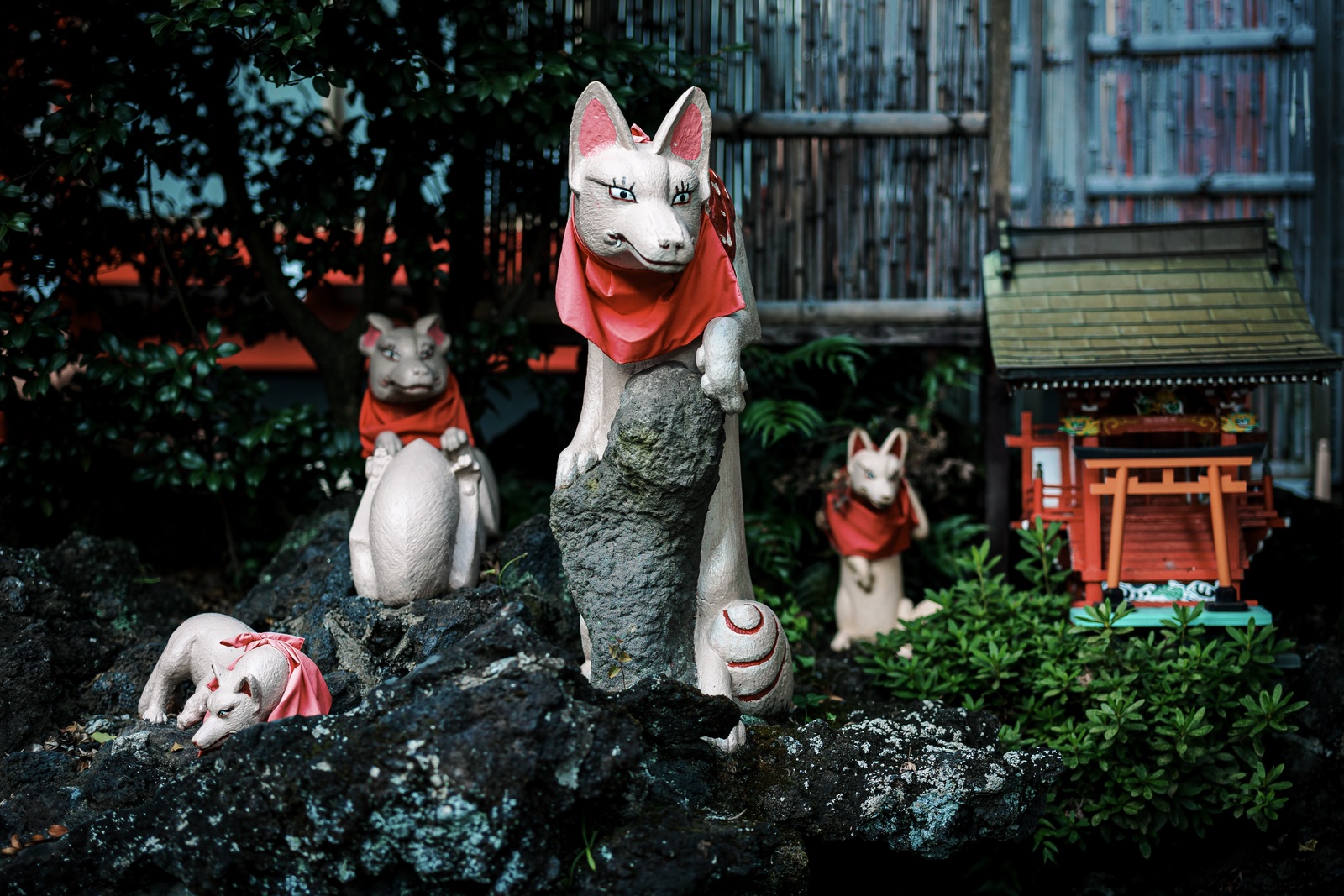 White fox statue leaning over dark volcanic rock, with several other foxes and a small shrine behind it at Keihin Fushimi Inari Shrine.