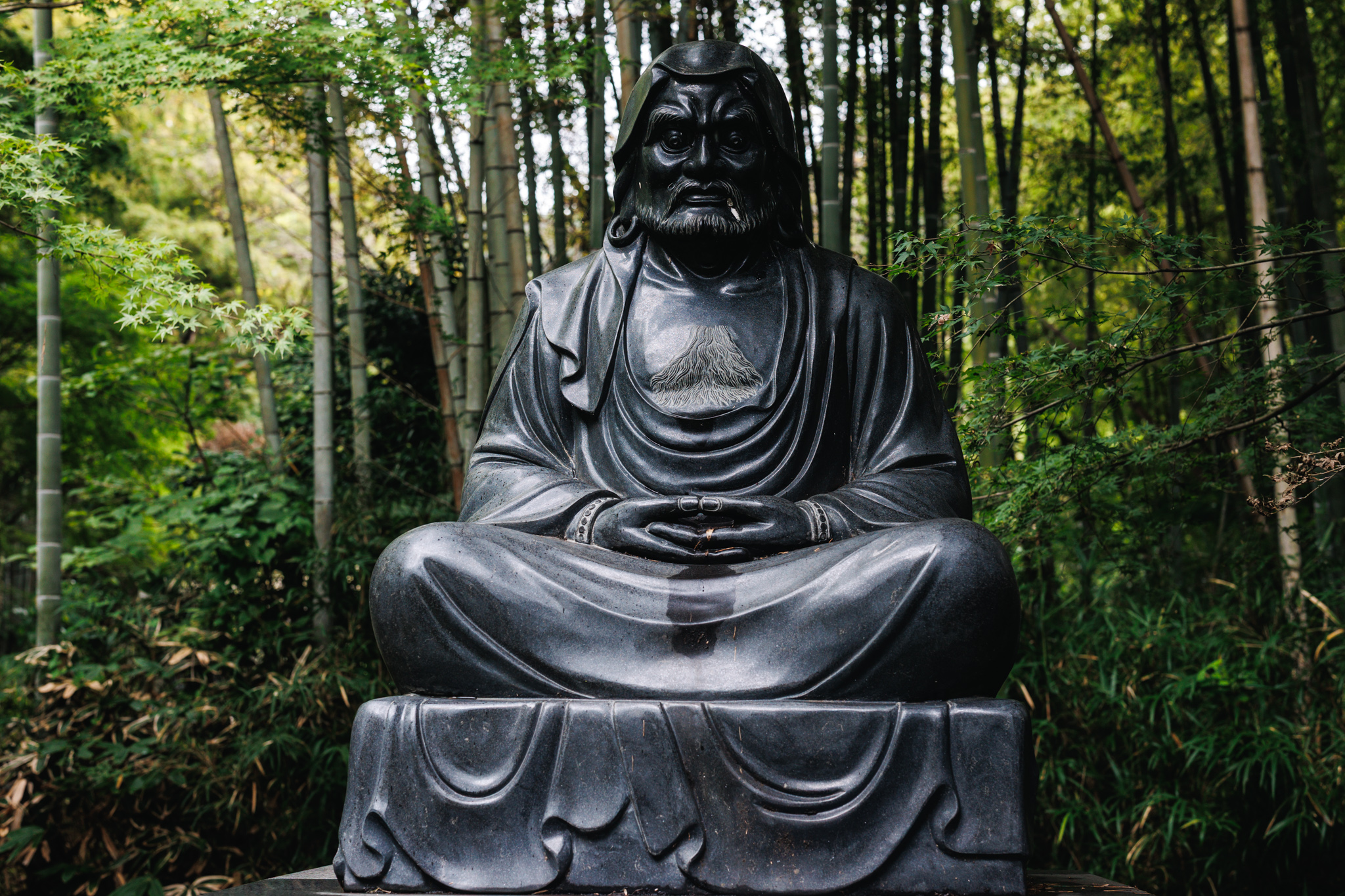 A seated Daruma statue on the path toward Hanzōbō at Kenchōji Temple, backed by bamboo.