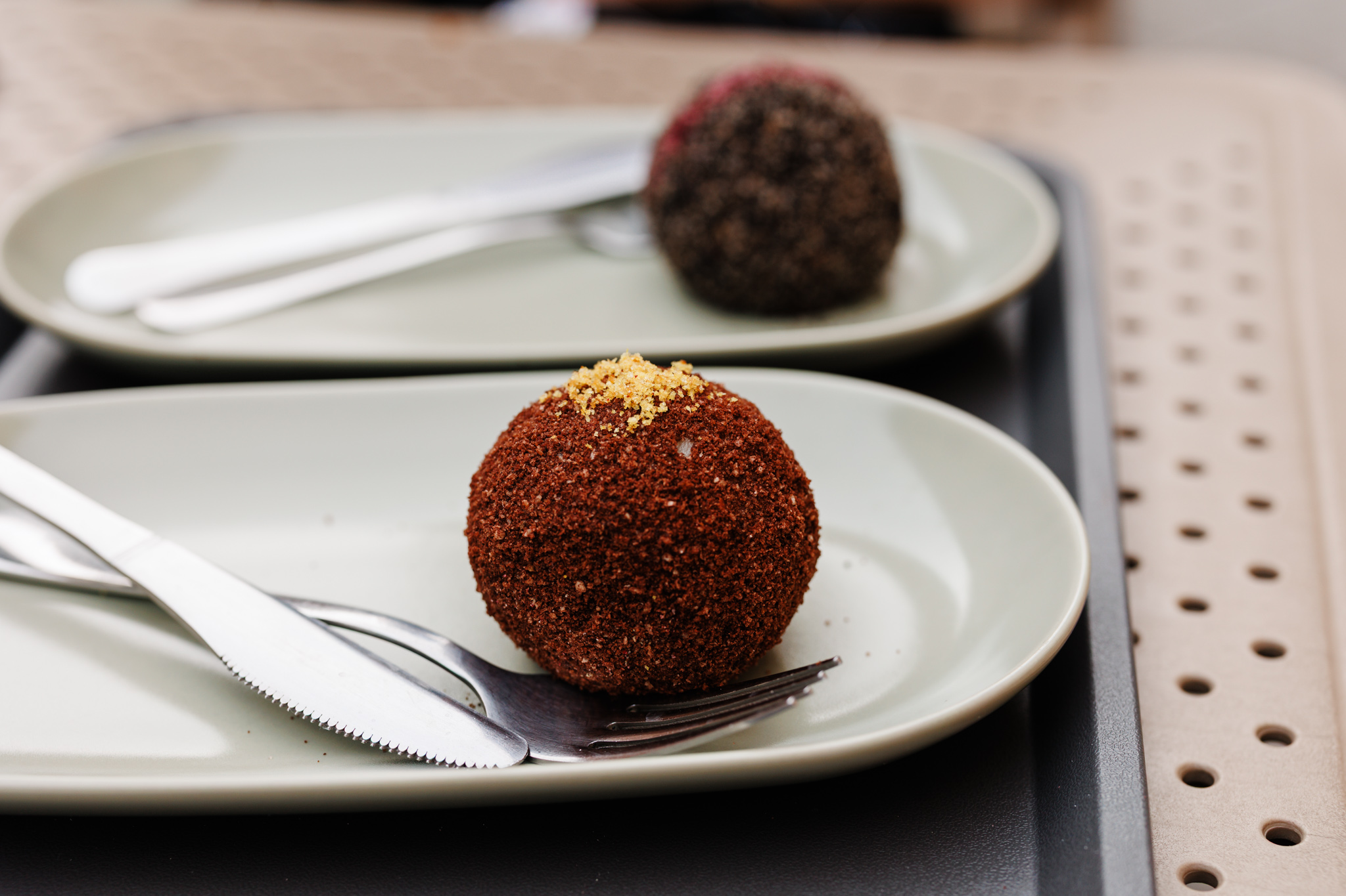 A chocolate-coated knedla with a small golden topping on a pale plate at Ferdinand knedle in Novi Sad, with another knedla blurred behind it.