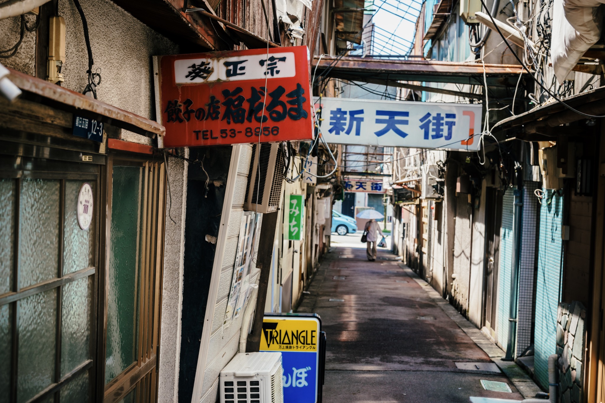 Narrow Kofu Shintengai alley with old shopfronts, dense wiring, the bare frame of its former arcade roof and a pedestrian beneath the Shintengai sign.
