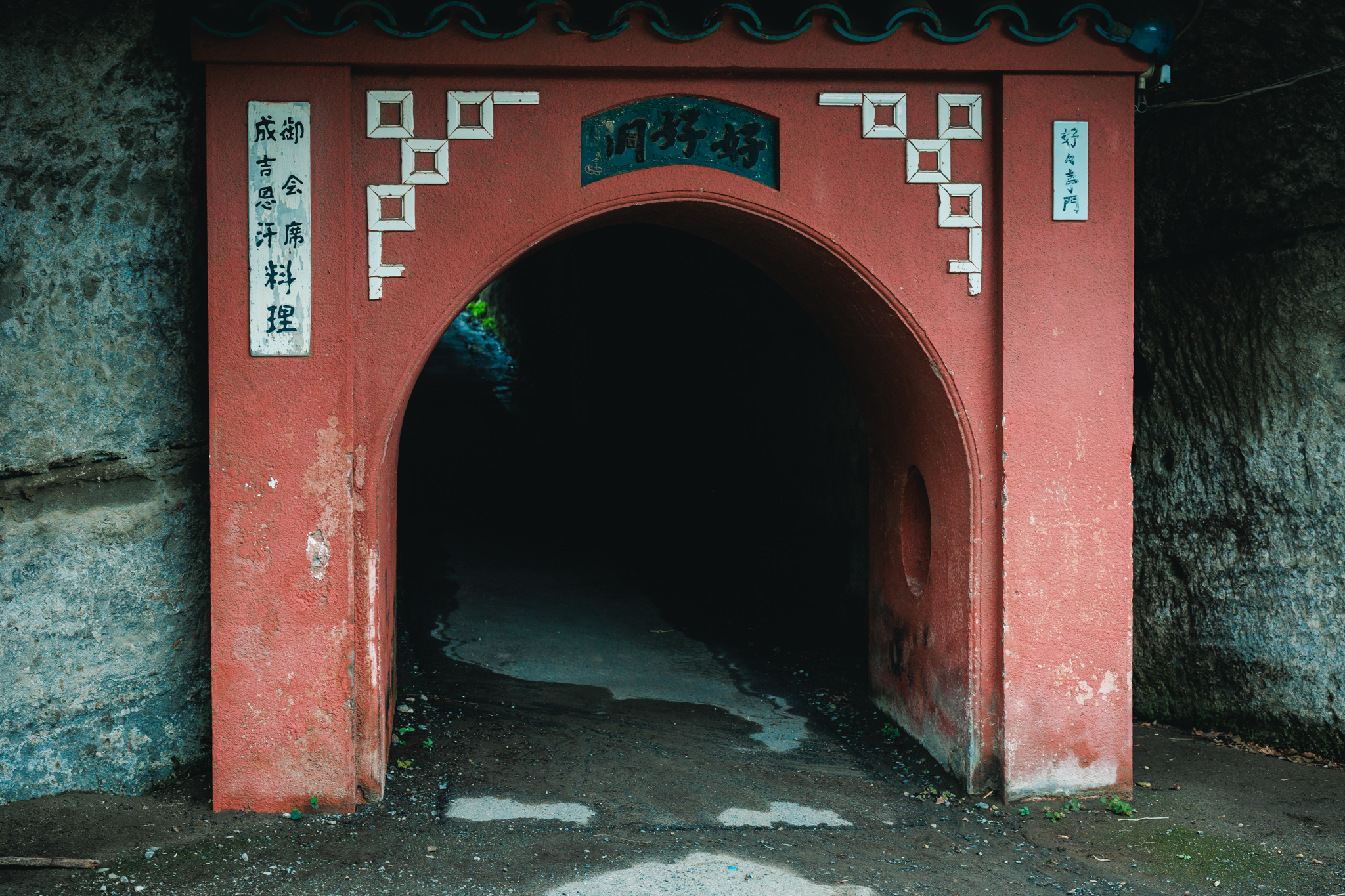 Red gateway of Kōkōdō tunnel in Kamakura, with faded food signs and a dark pedestrian passage beneath the rock.