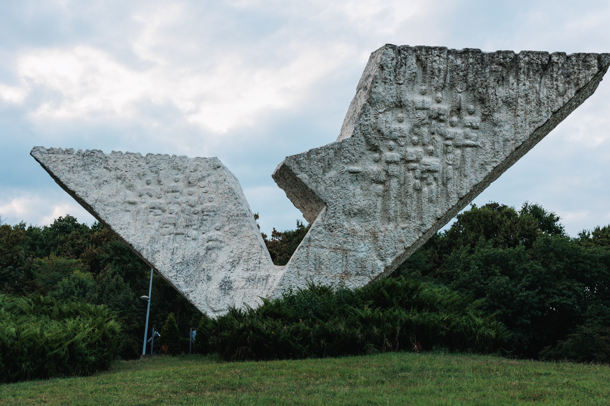 Two massive white concrete wings forming a V above a hedge, their inner faces covered with small relief figures, under a cloudy sky
