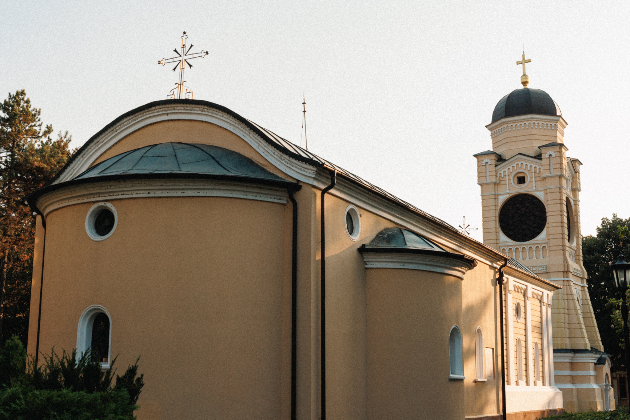 The rounded apse and a side bay of the Old Church, ochre walls under a dark metal roof with no dome, and the cream bell tower with a large round grilled opening beyond