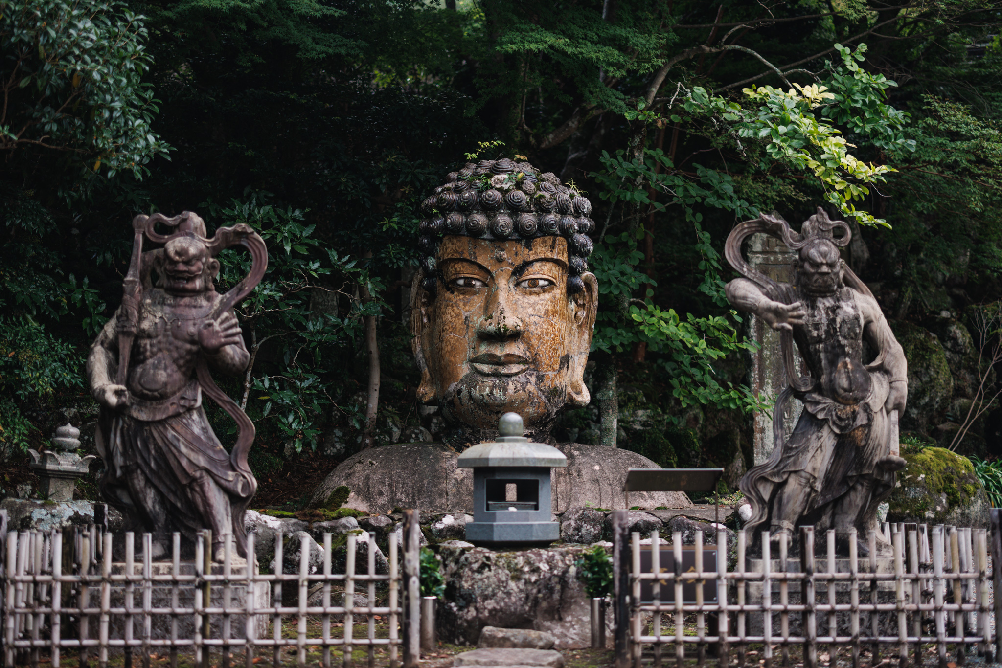 A large ceramic Buddha head with tightly curled hair on a broad, rounded base, behind a bamboo fence and a stone lantern, with a dark guardian figure standing on either side and trees behind