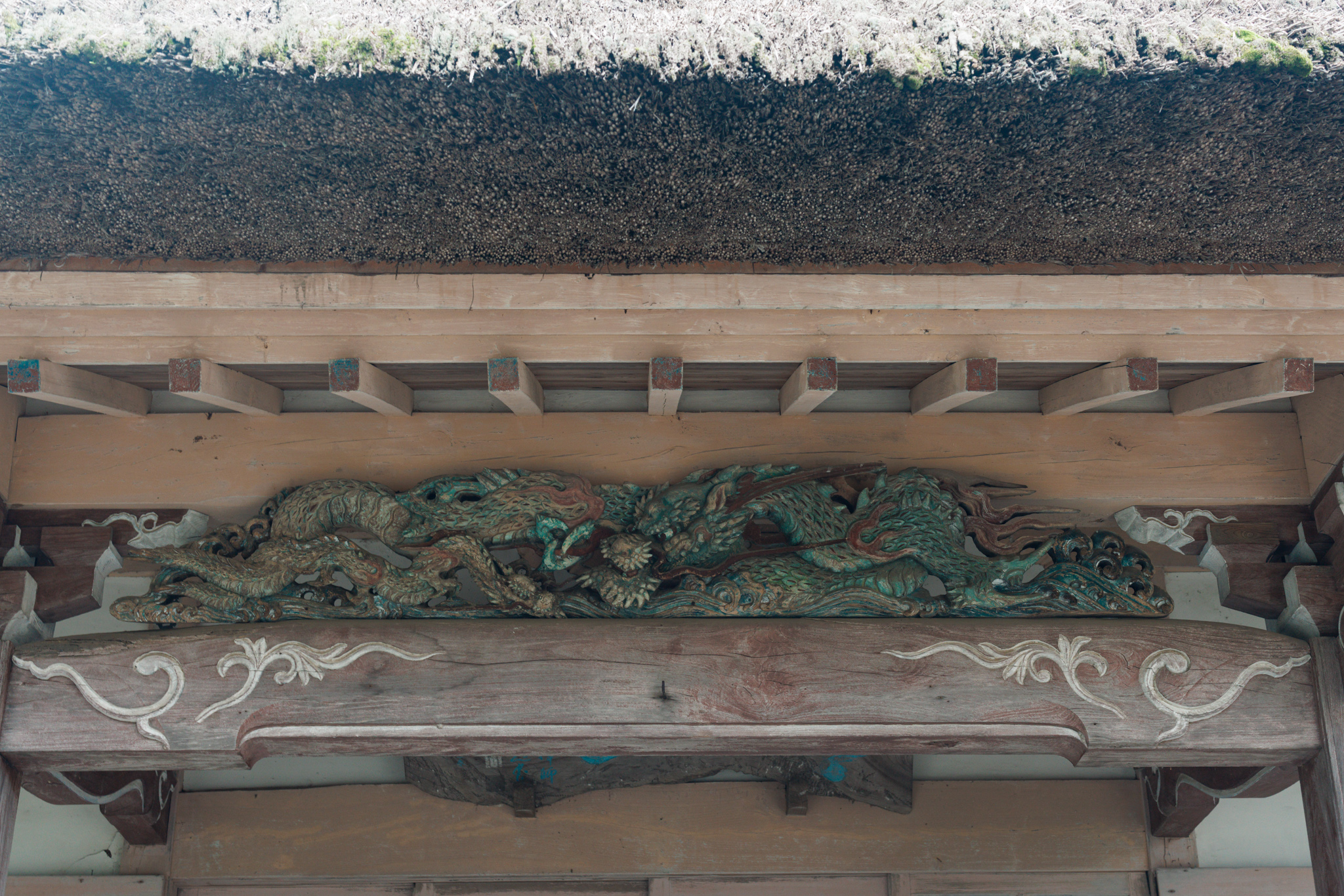 The underside of a thick thatched eave, with a green-painted carving of dragons along a beam above a board decorated with white scrolls