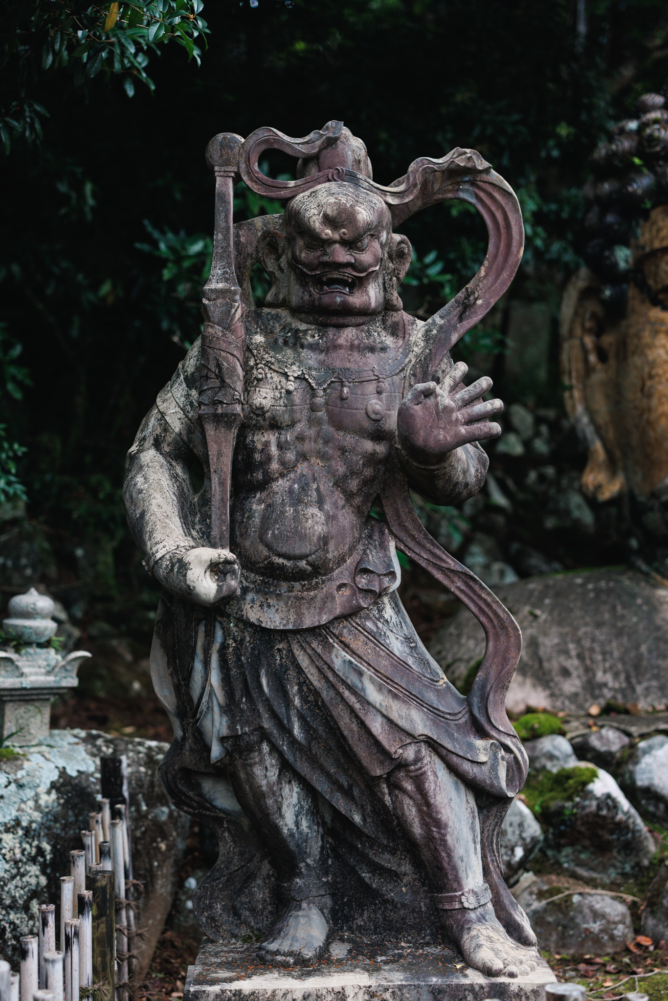 A dark guardian figure with an open mouth and a fierce expression, holding a long staff, with a sash curling behind its head