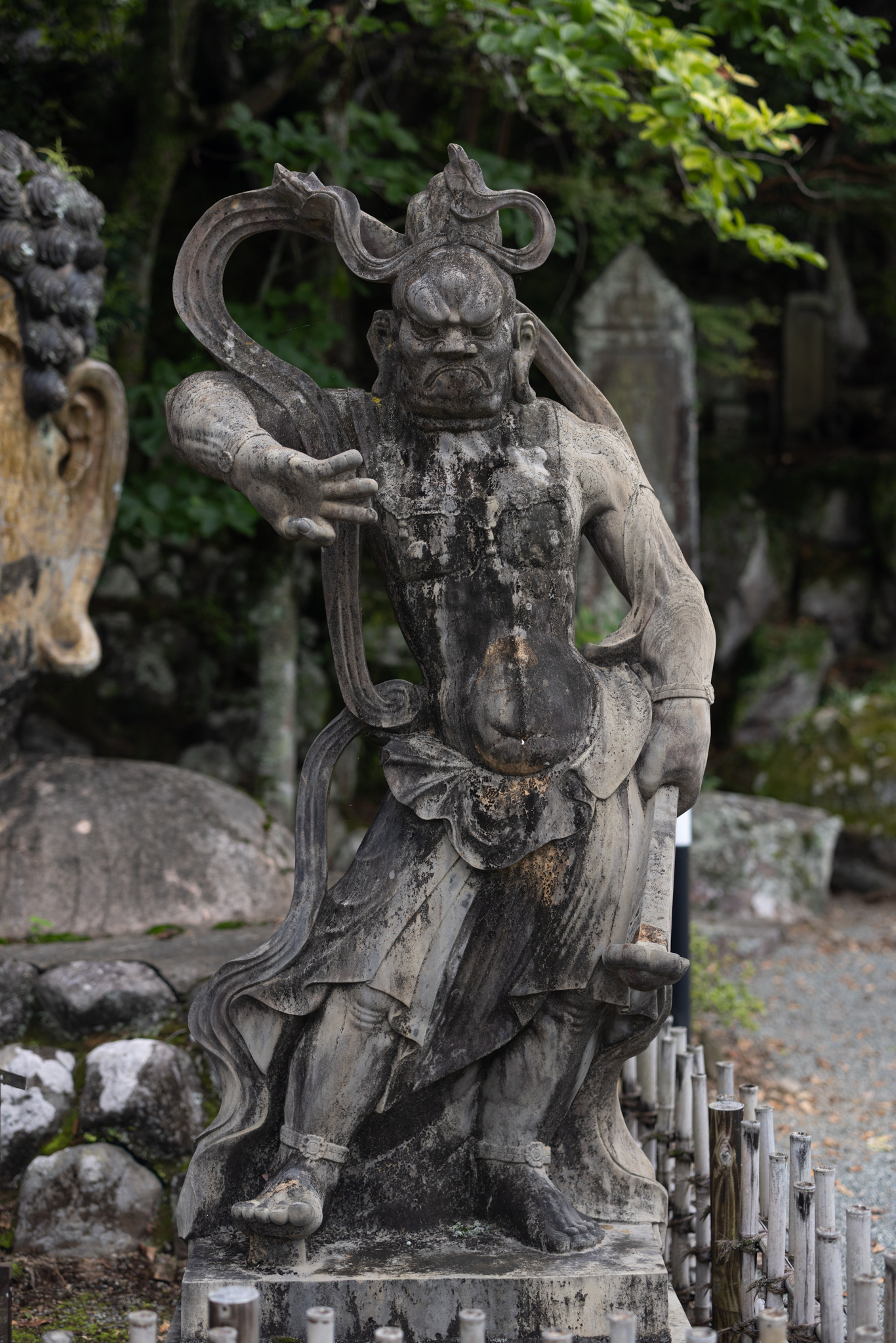 A second guardian figure with a closed mouth, one arm raised across its body, standing beside the edge of the Buddha head