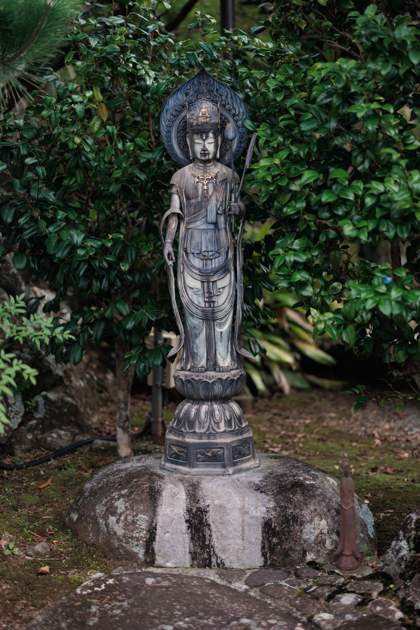 A slender standing figure with a crown and a halo on a lotus pedestal, holding a long stem, set on a rock among shrubs