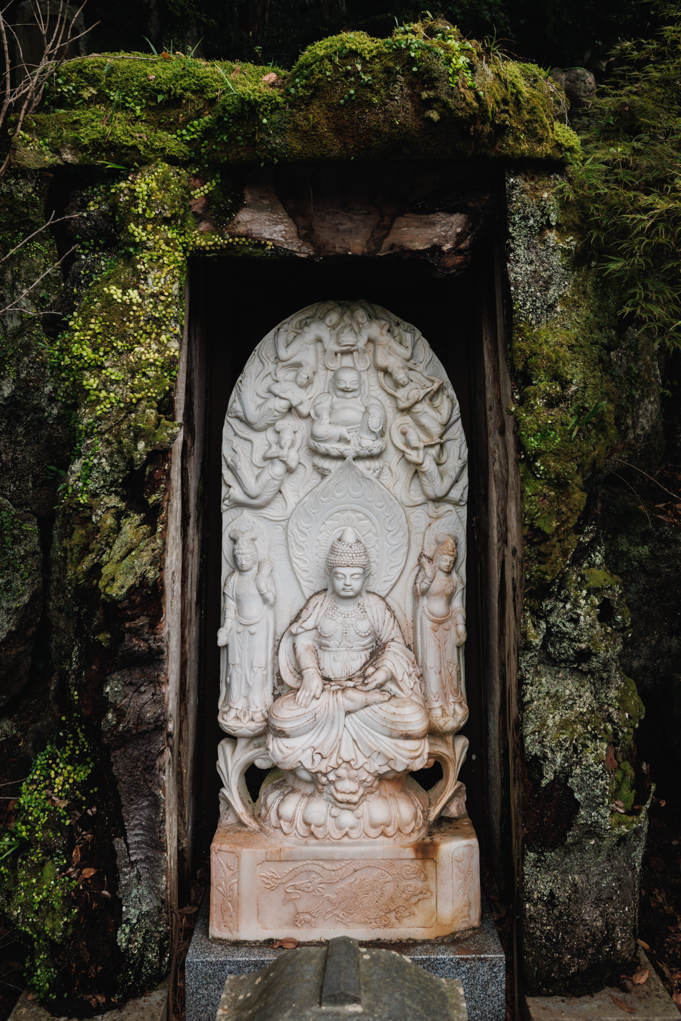 A white relief of a seated figure with attendants and figures above, set in a niche under a heavy stone roof covered in moss