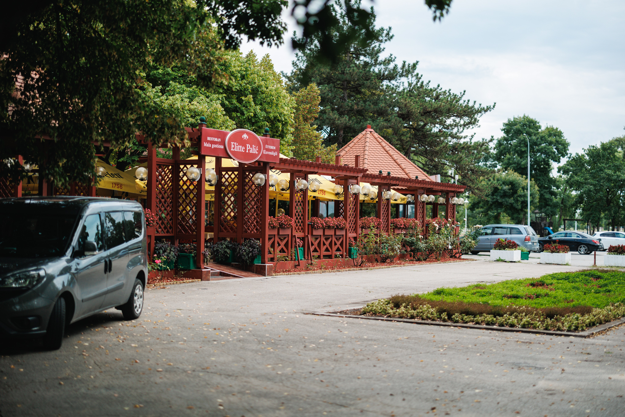 A red pergola-style restaurant front with a sign in Serbian and Hungarian and yellow parasols behind it