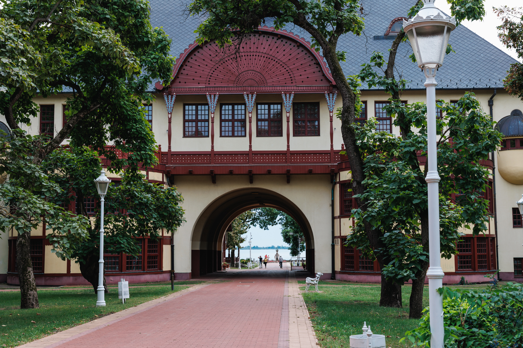 The Grand Terrace at Palić seen from the park, a carved dark-red wooden gable above an arched passage with the lake visible at the far end