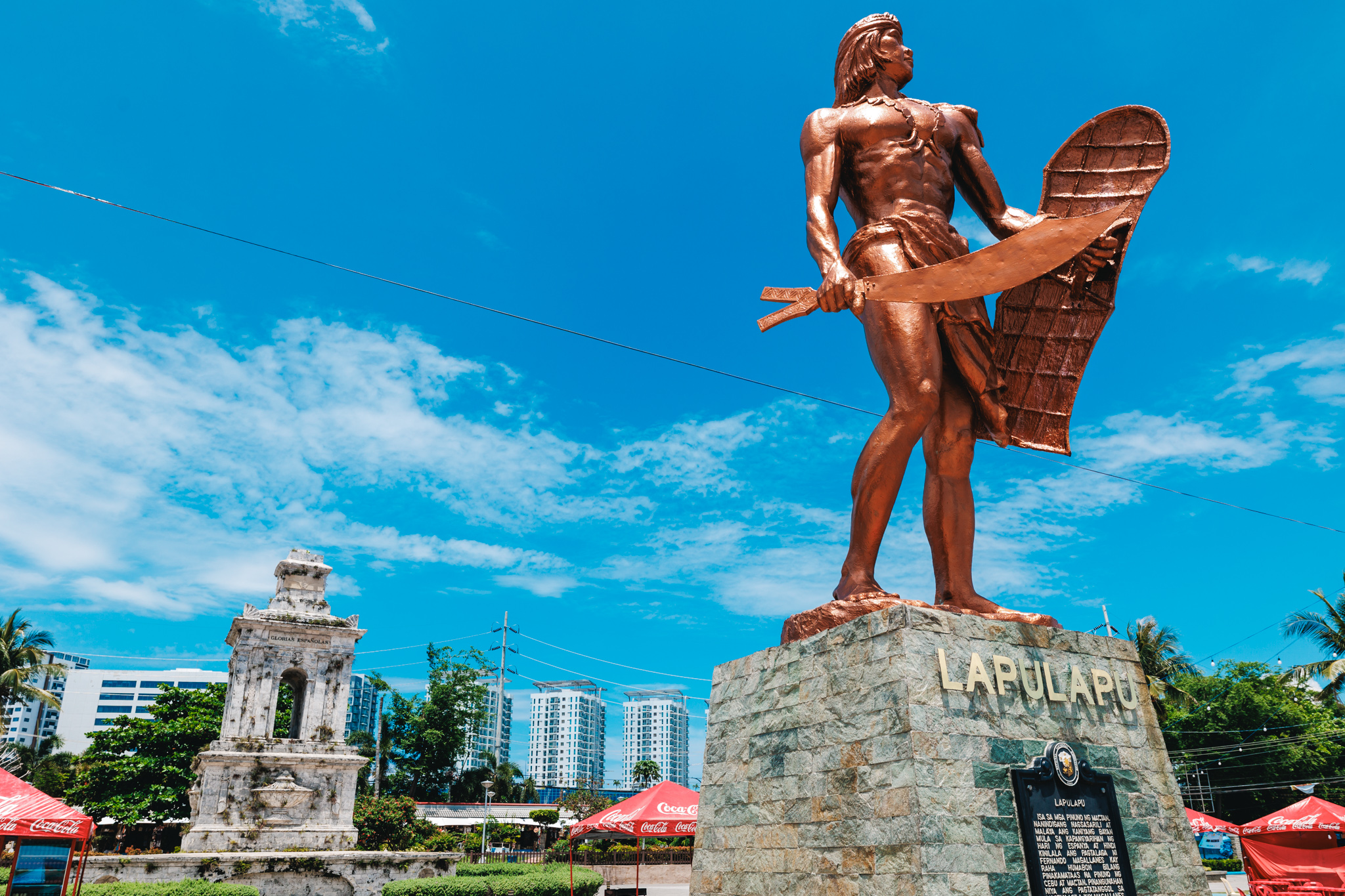A copper-coloured statue of Lapulapu holding a curved blade and a shield on a stone pedestal, with the white stone Magellan obelisk, its top missing, behind it and red Coca-Cola umbrellas below