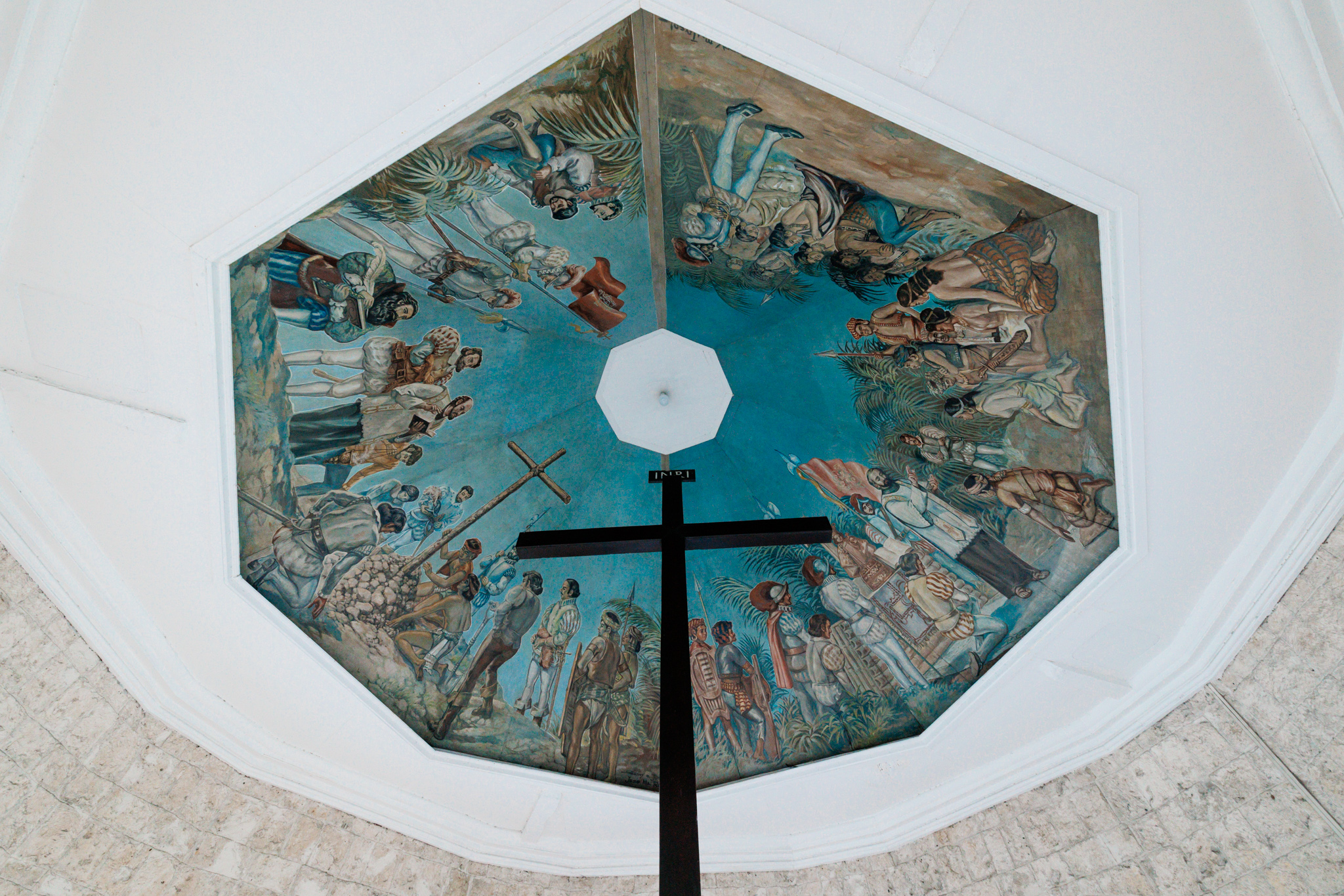 Looking up past the top of a dark wooden cross marked INRI at an eight-sided ceiling painting on a blue ground: men hauling a cross upright on one side, a priest at an altar before a crowd on the other