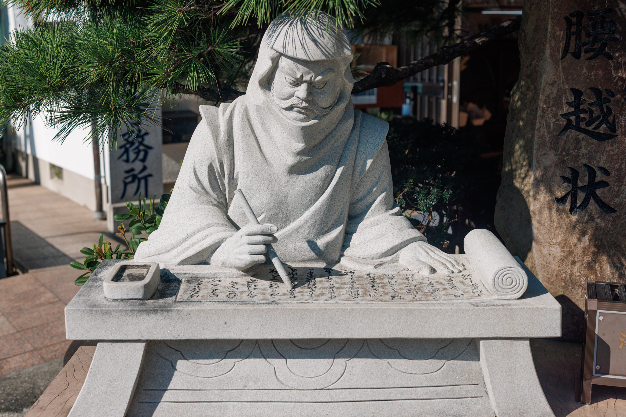 A grey stone statue of Benkei in a hood, seated at a stone desk with a brush in his hand and a scroll unrolled in front of him, under pine branches, beside a tall stone carved with the name of the Koshigoe letter