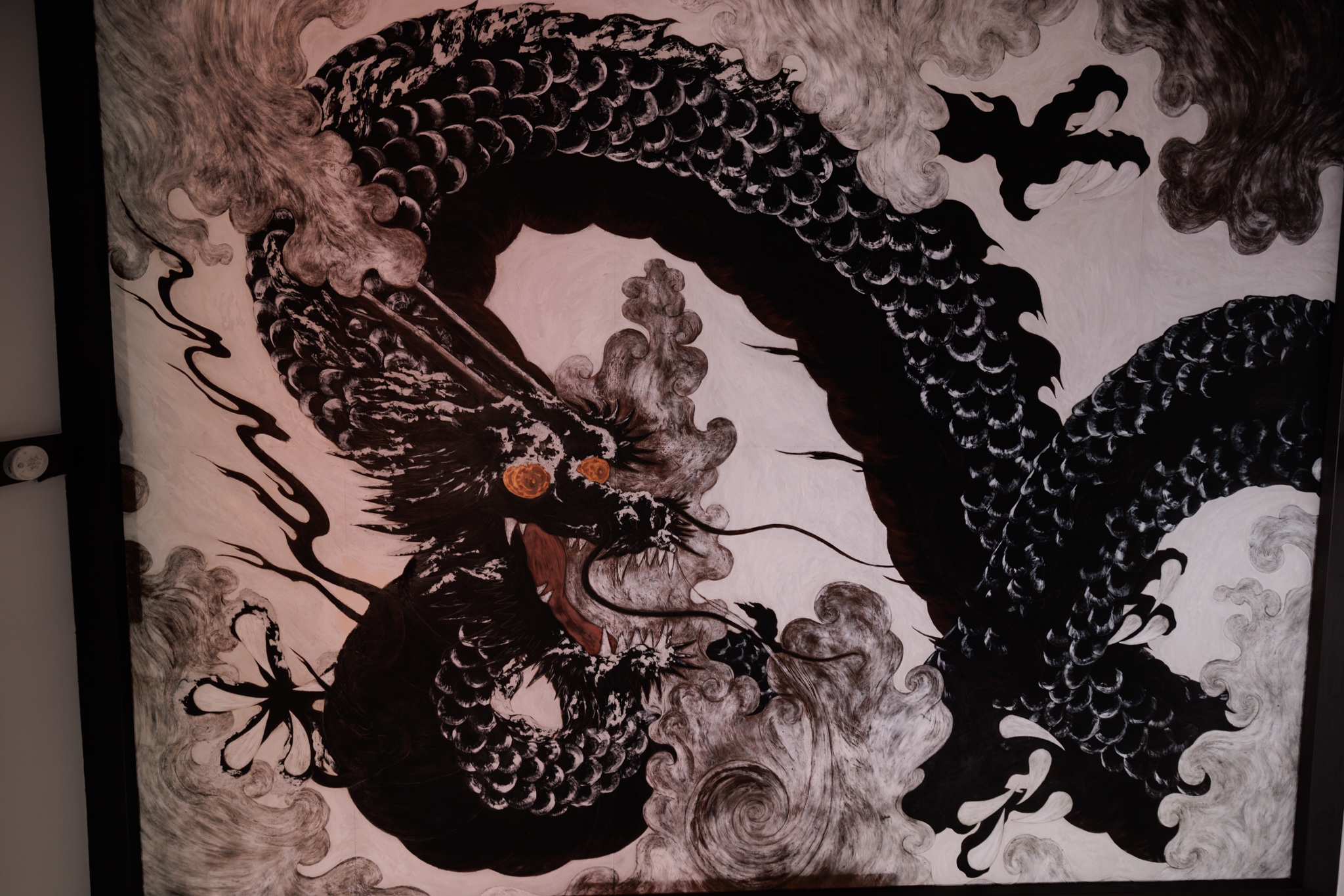 A black dragon with orange eyes painted on a pale ceiling panel among swirling clouds