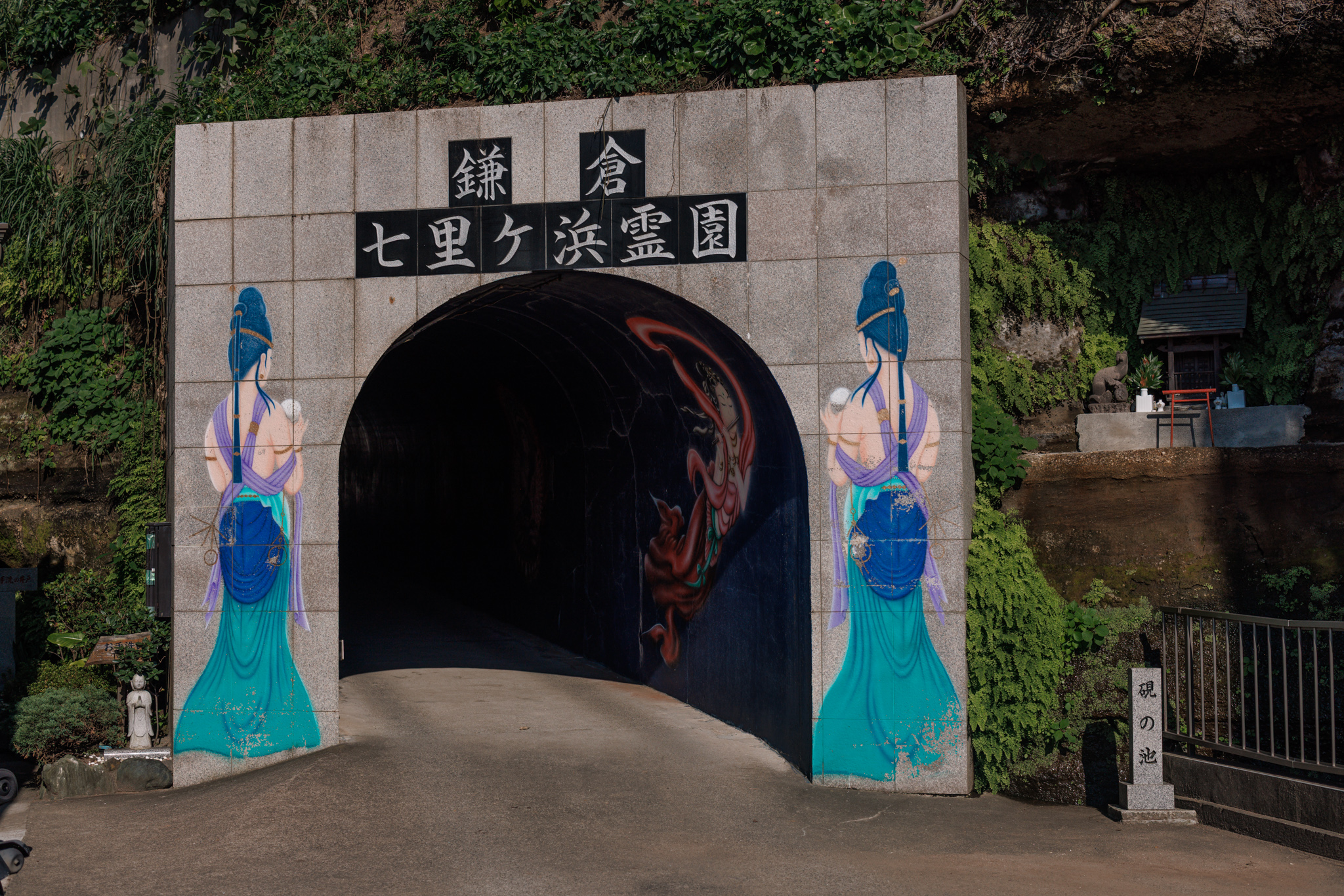 An arched tunnel entrance under a sign reading Kamakura Shichirigahama Cemetery, with two women in flowing blue and green robes painted on either side and a small stone marker by a railing on the right