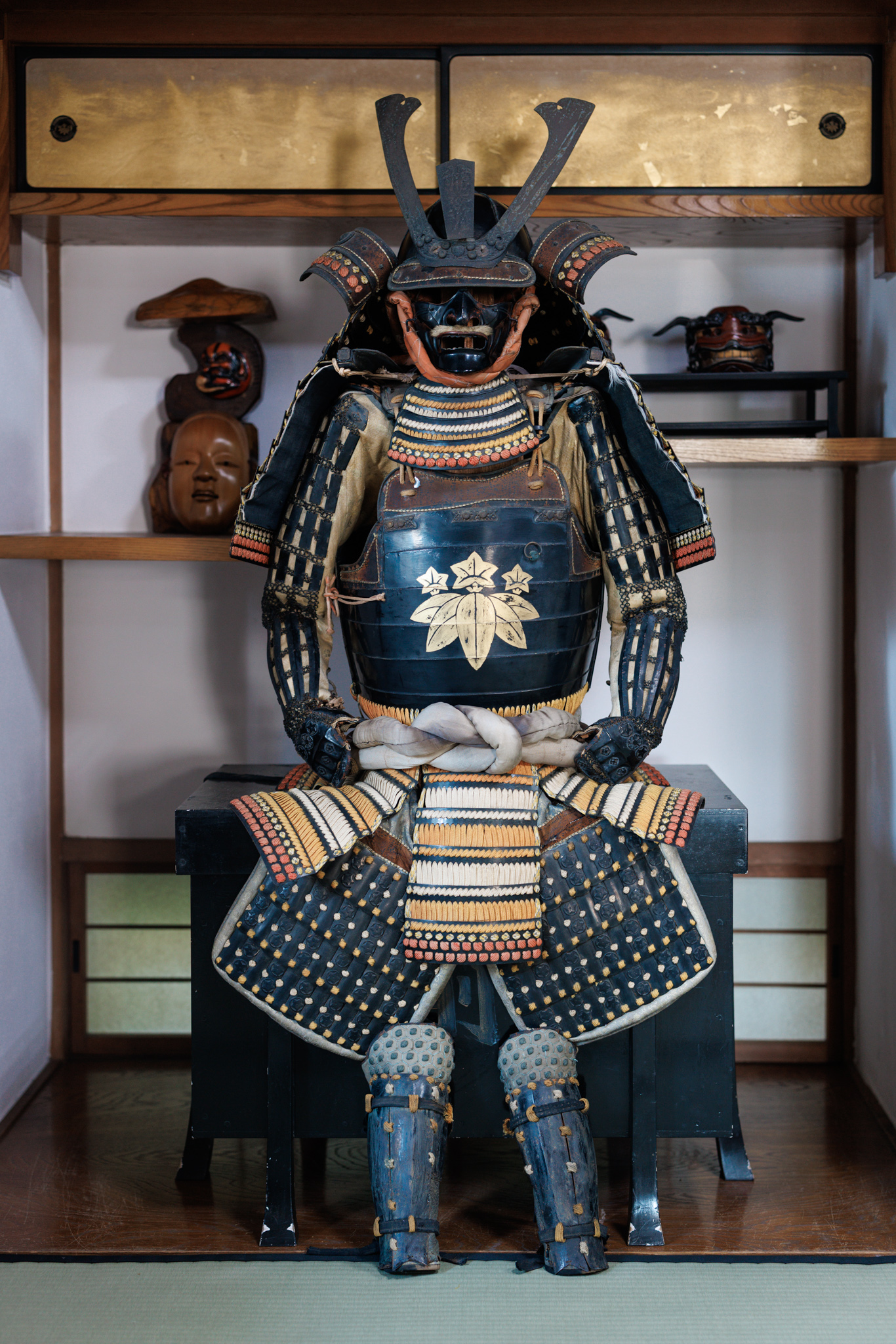A suit of black and gold armour with a horned helmet, displayed seated in an alcove, with a gold bamboo-and-gentian crest on the chest