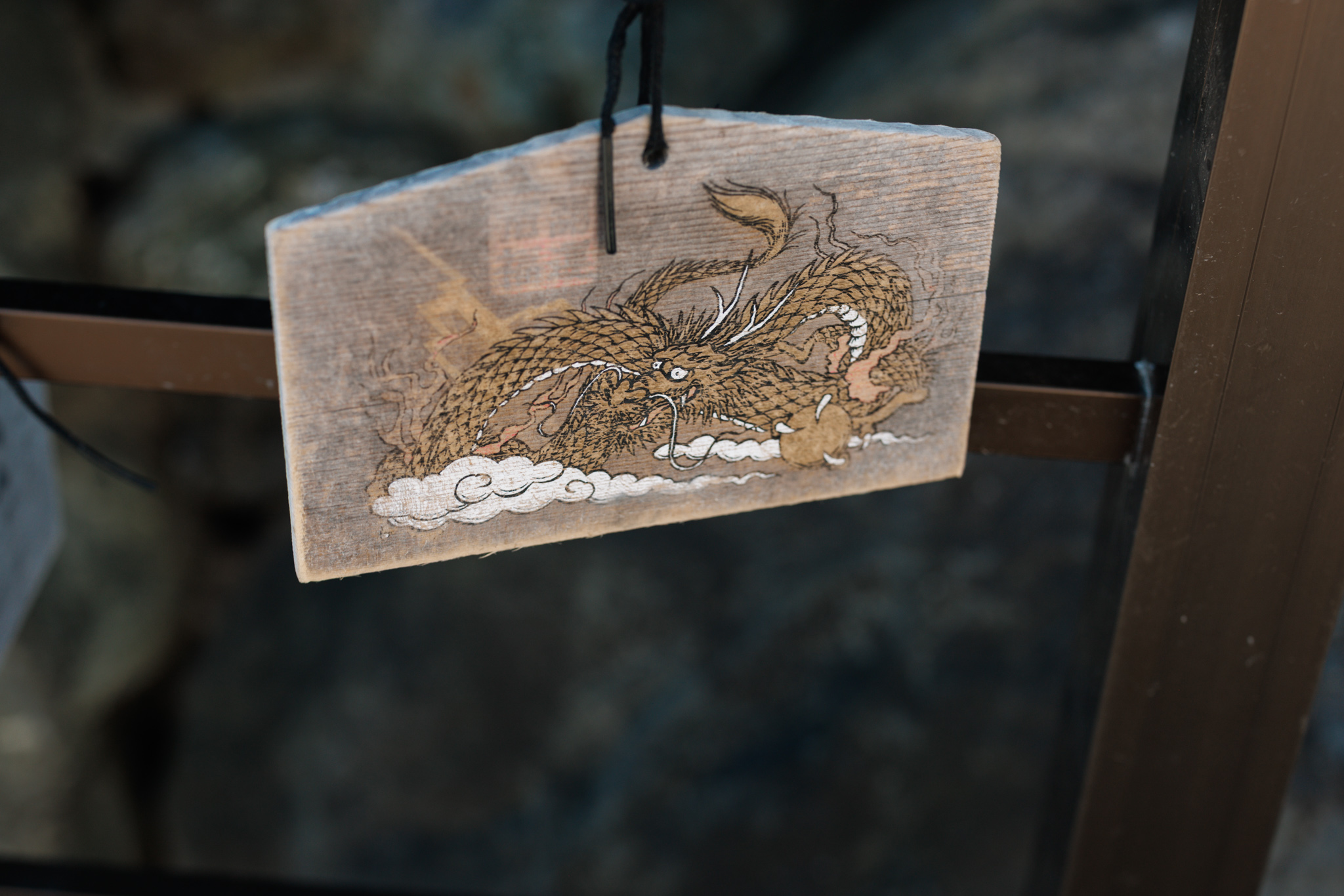 A weathered wooden ema plaque painted with a gold dragon among clouds, hanging from a dark cord