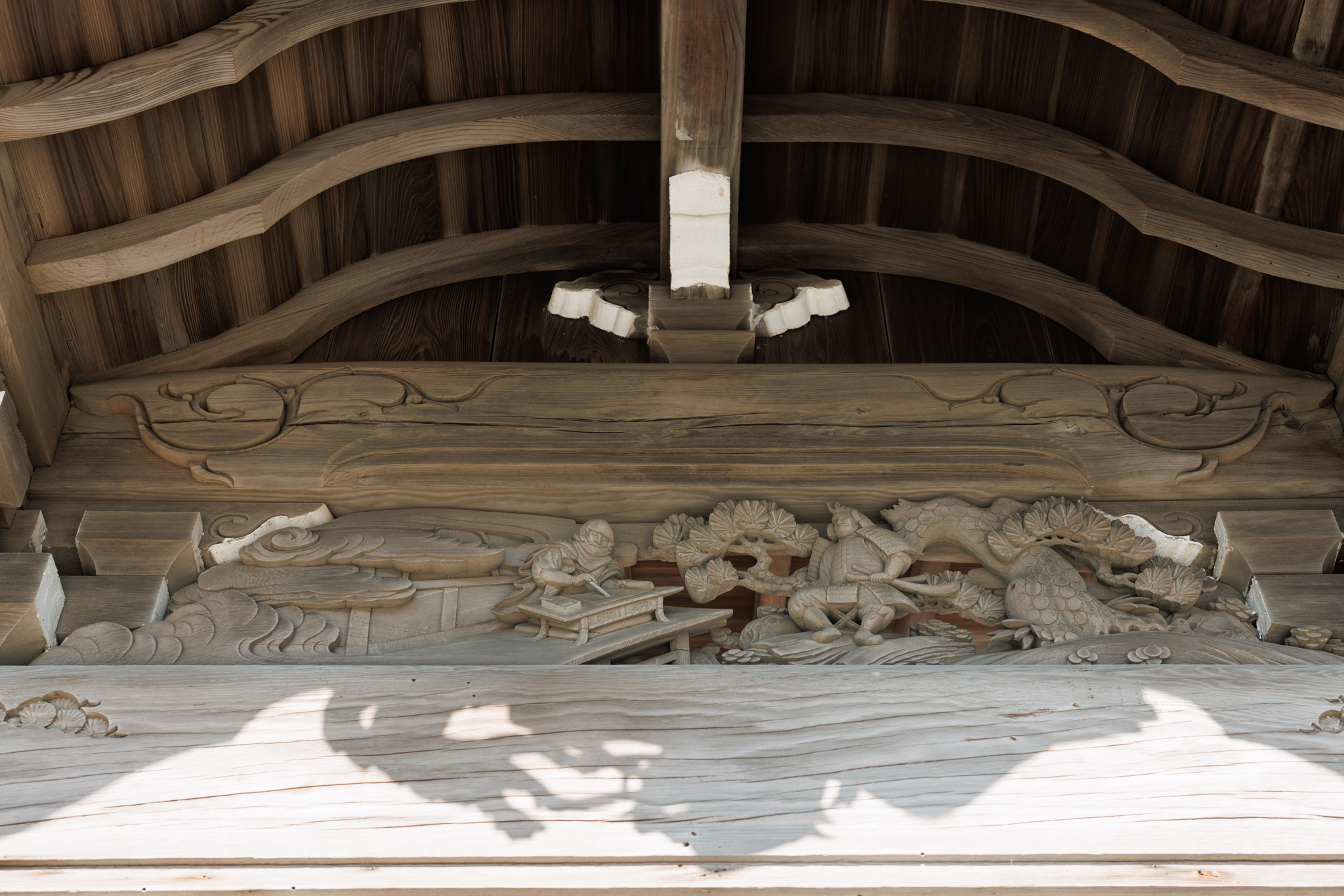 A weathered wooden carving under the eaves of the main hall: a hooded figure writing with a brush at a low desk on the left, and a seated warrior in armour among pine branches on the right