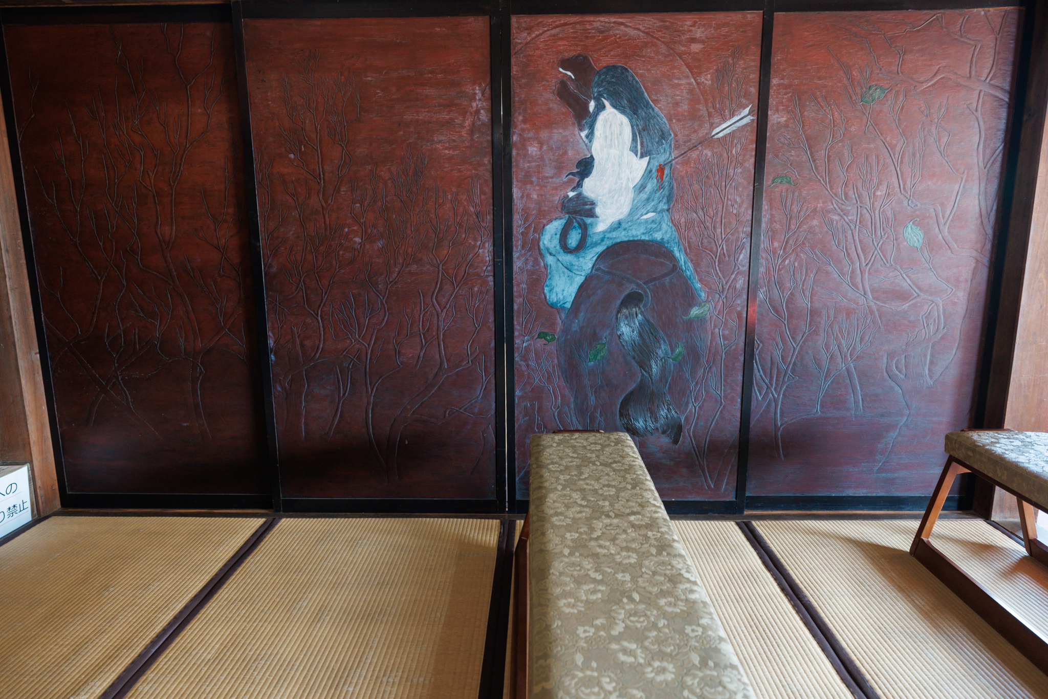 Dark red sliding doors carved with bare branches, the third painted with a white-hooded figure and an arrow, seen from a tatami room with a bench in front