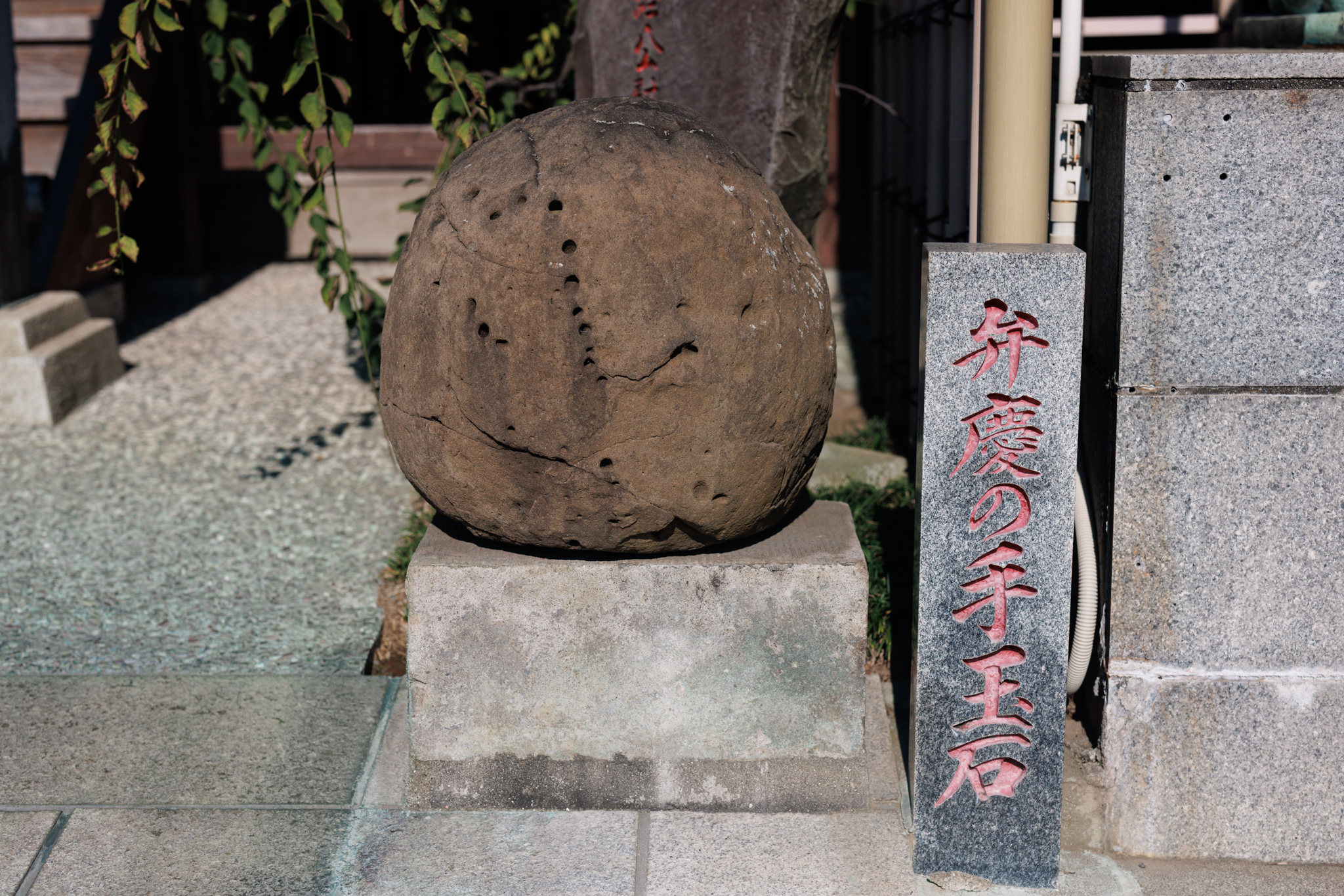 A large round stone pitted with small holes, set on a stone base, beside a stone marker naming it as Benkei’s juggling stone