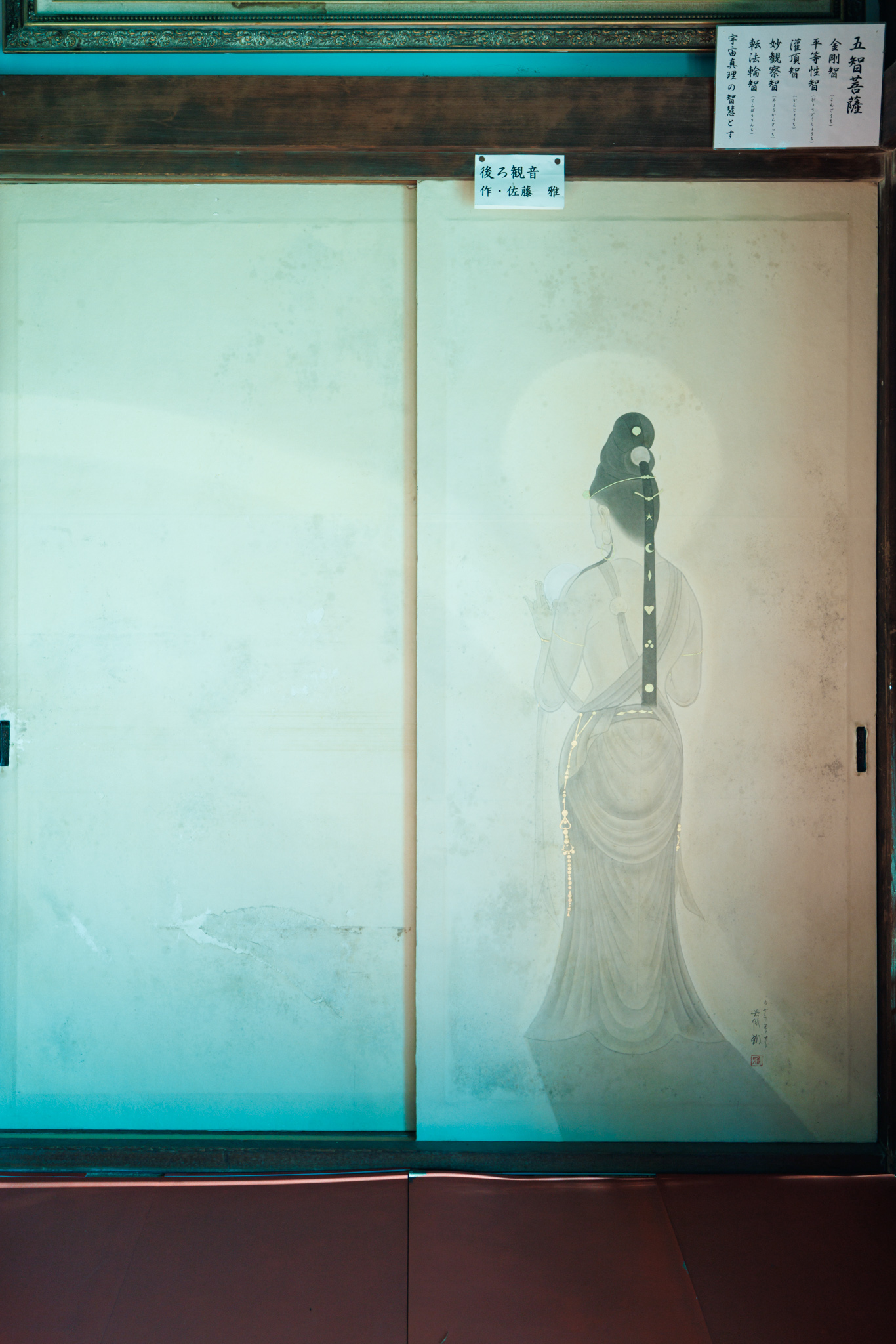 A pale sliding door painted with a Kannon seen from behind, with long dark hair, a halo and flowing robes, under a small title label