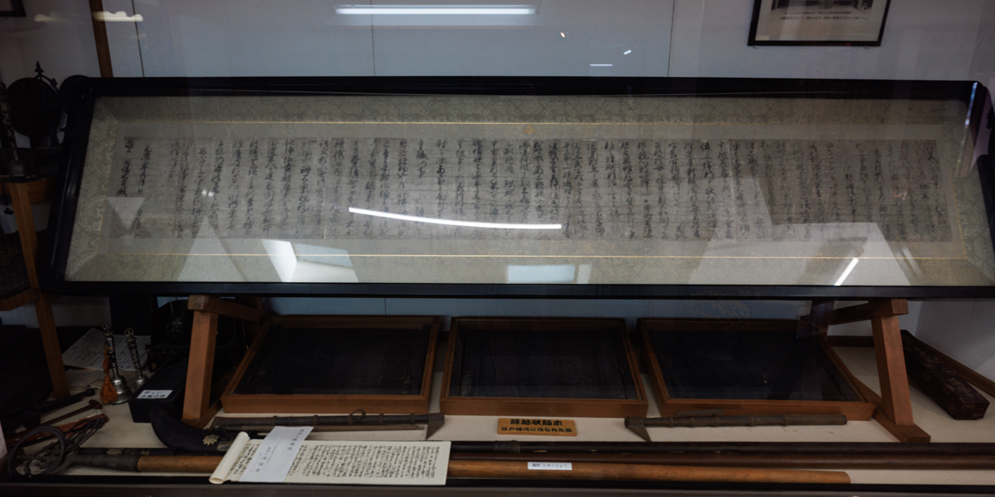 A long handwritten letter mounted under glass in a display case, with three wooden trays of woodblocks, long staffs and a printed copy of the letter on the shelf below