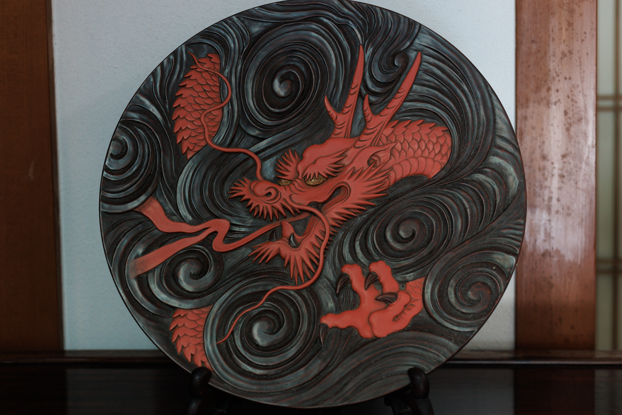 A round black panel carved with swirling waves and a red dragon with outstretched claws, propped on a stand