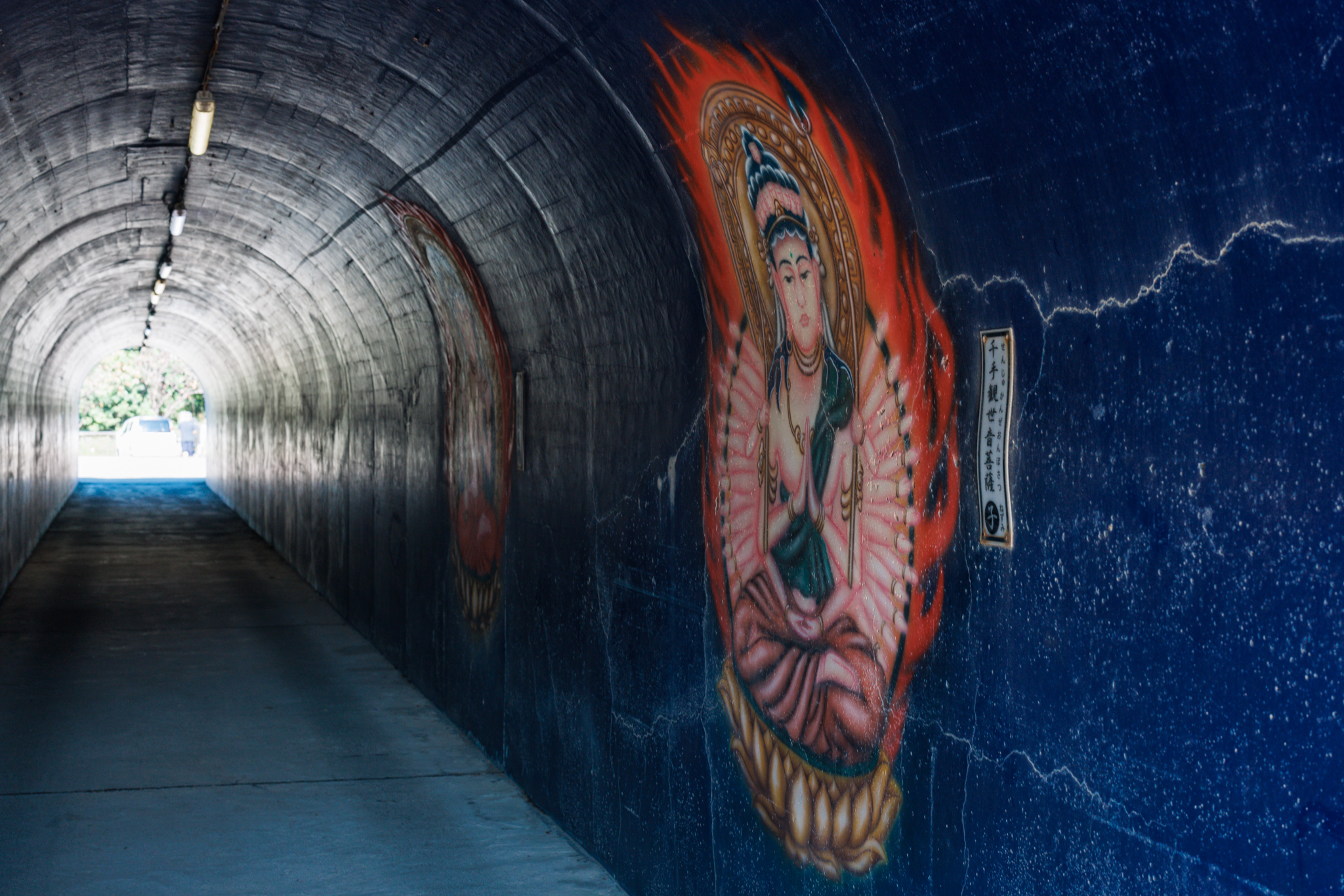 Inside a tunnel painted dark blue, a many-armed Kannon surrounded by orange flames on the right-hand wall, with daylight at the far end