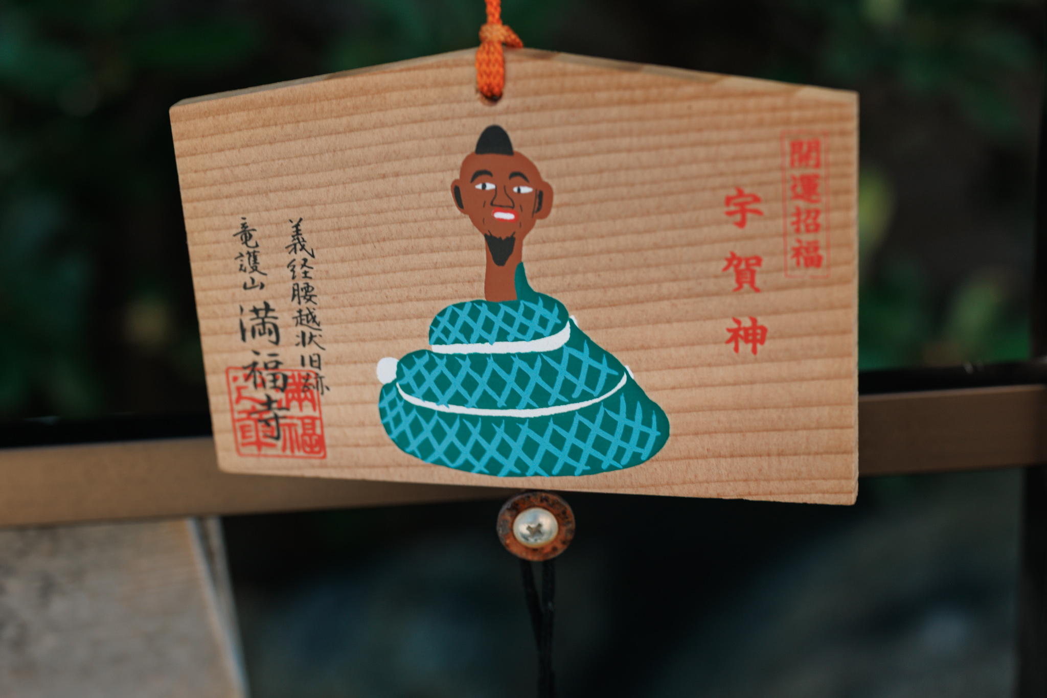 A wooden ema plaque painted with a bald human head on a coiled blue-green snake’s body, inscribed with the name Ugajin and the temple’s name