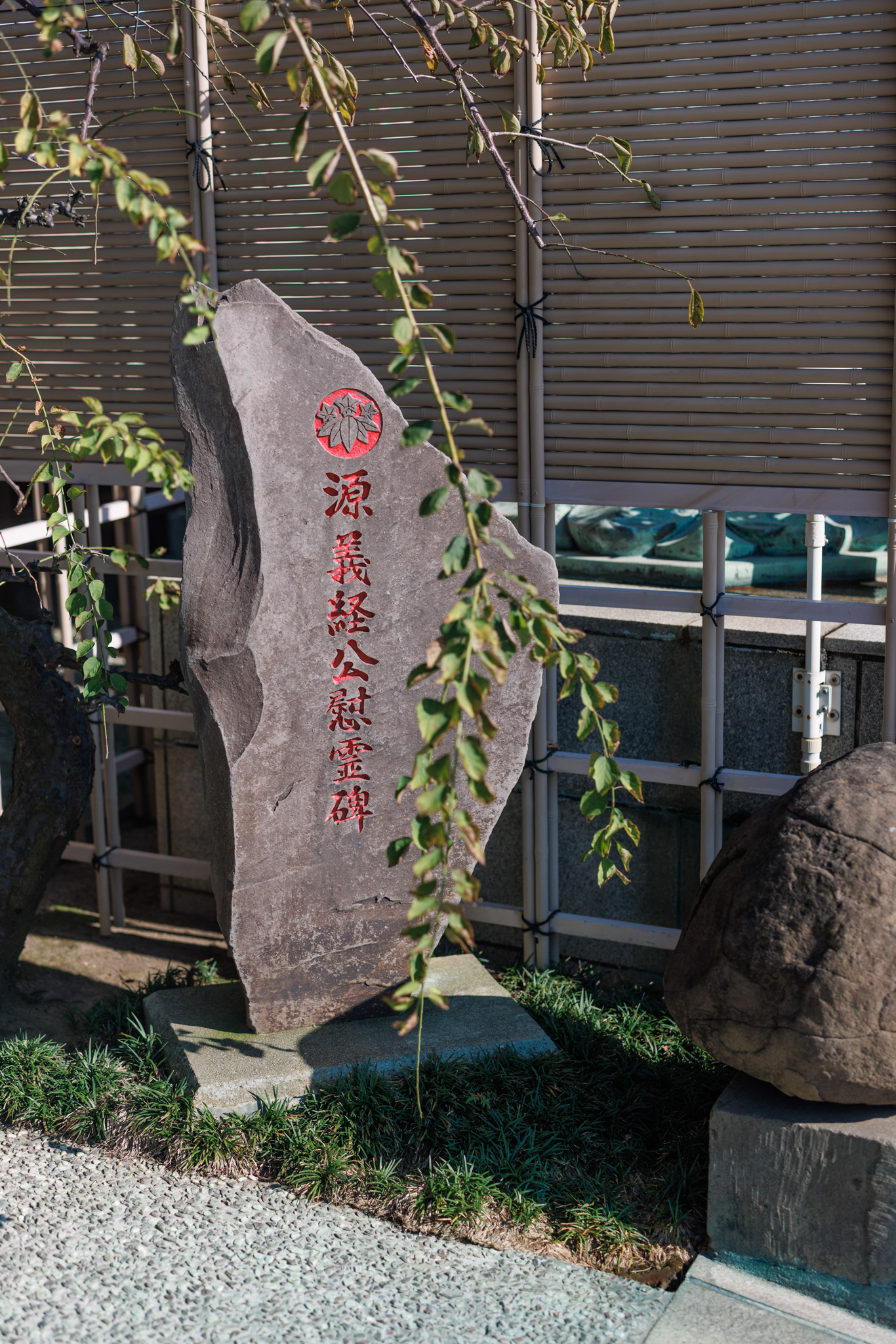 A tall, narrow memorial stone carved with red characters beneath a red bamboo-and-gentian crest, among shrubs and hanging branches