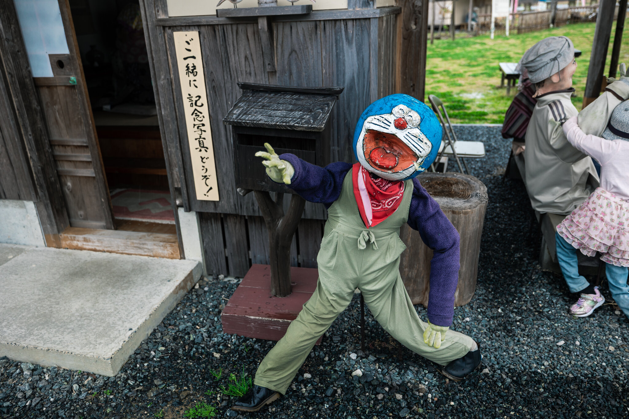 A Doraemon-like handmade scarecrow stands beside a sign inviting visitors to take a souvenir photograph.