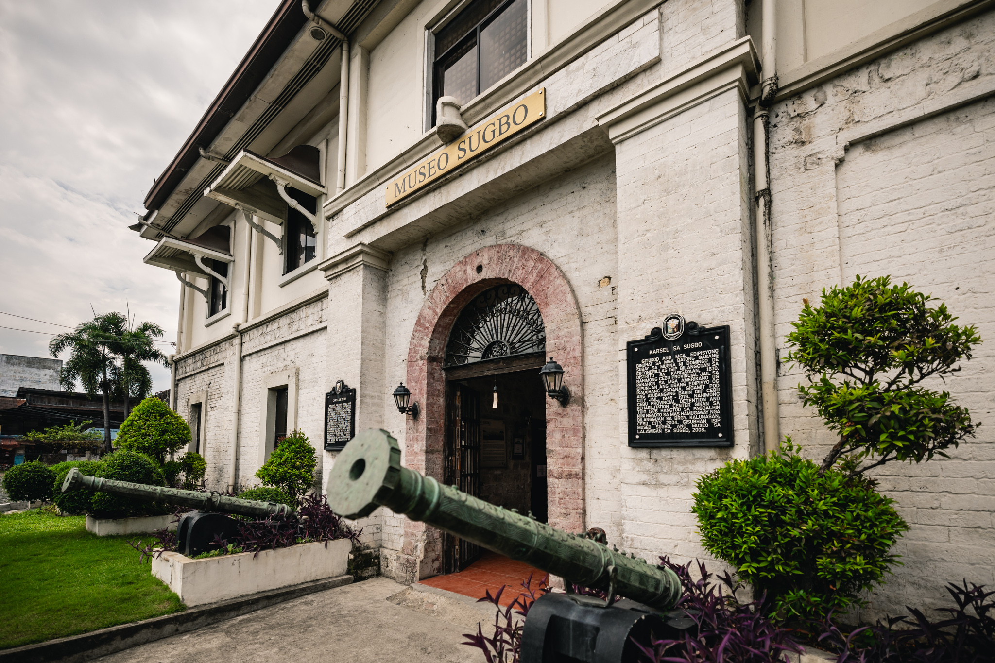 An angled view of the museum gate, with a long green-patinated gun in front and a black plaque headed KARSEL SA SUGBO on the white wall to the right