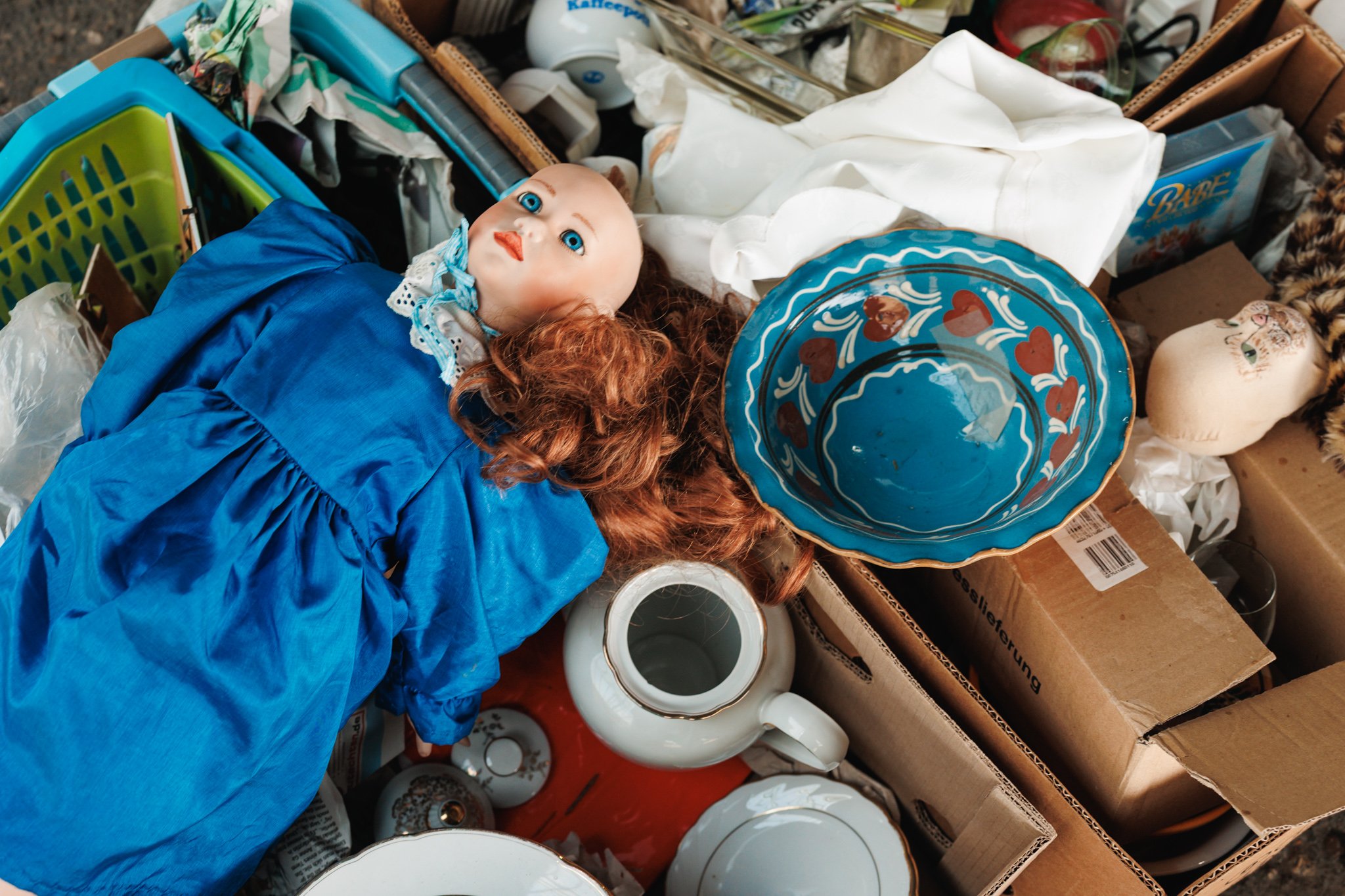 A doll in a blue dress lying beside a blue dish with red hearts, with a separate cloth doll head, white crockery and cardboard boxes at Najlon Market.