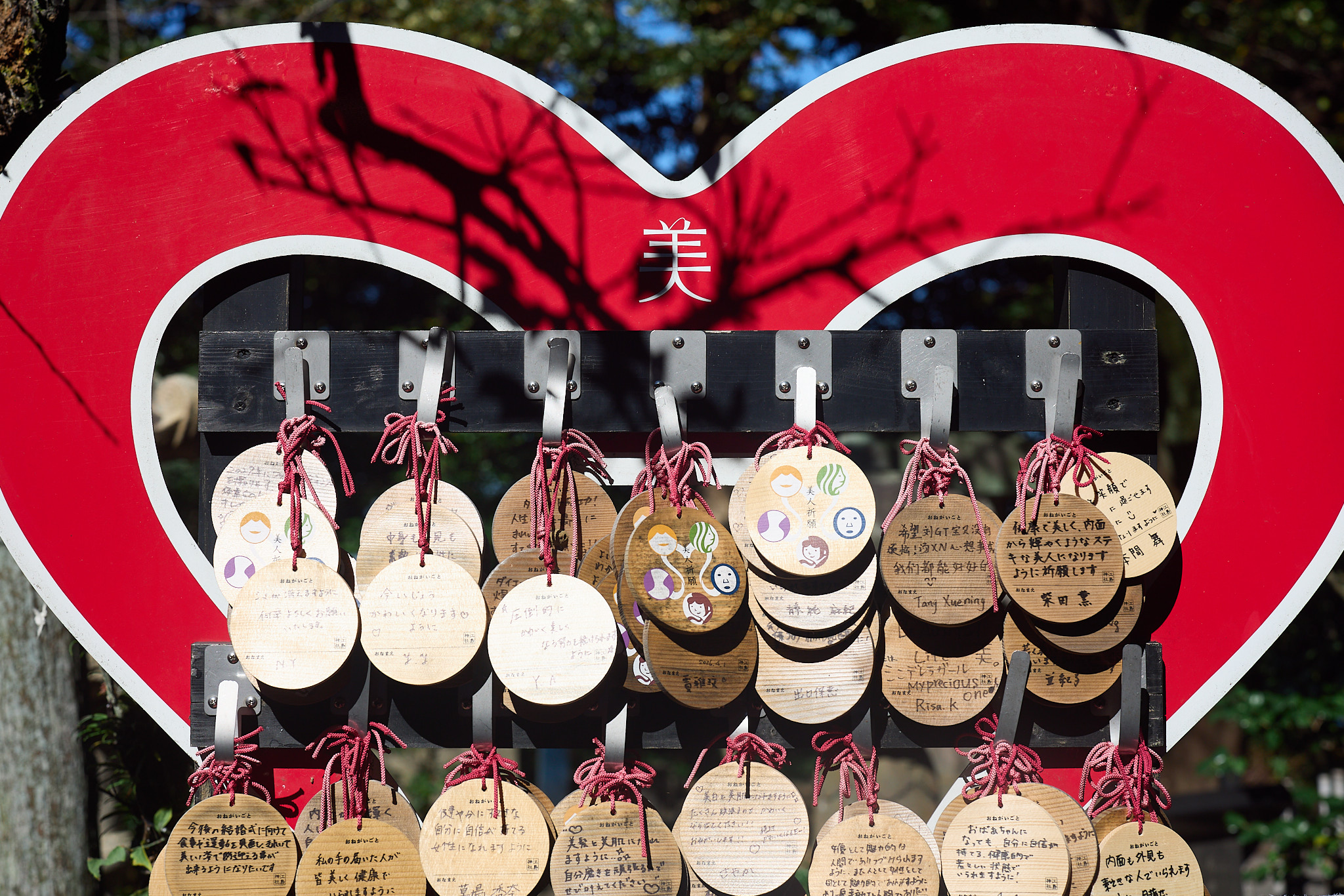 Round wooden prayer plaques with pink cords hanging inside a large red heart-shaped frame marked with the character 美