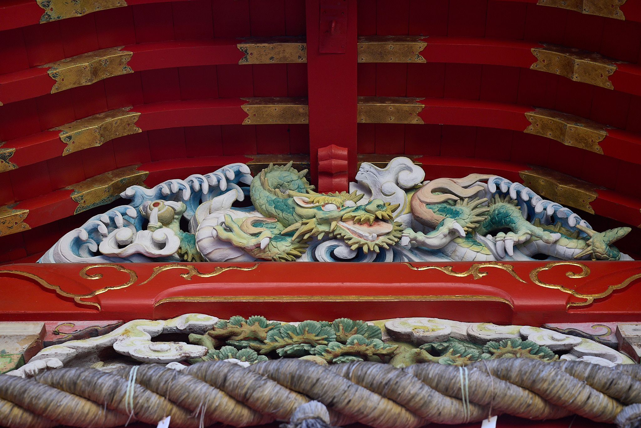 Painted carvings of green dragons among blue and white waves under red rafters, above a thick sacred straw rope