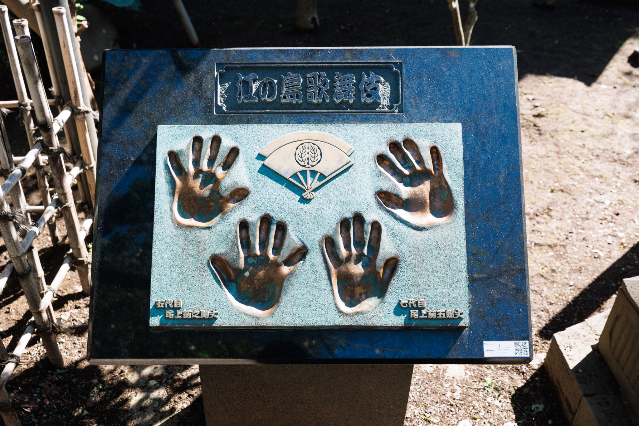A dark blue stone stand with a green bronze plaque bearing four handprints around a fan crest, the title “江の島歌舞伎” above and an actor's name in Japanese under each pair