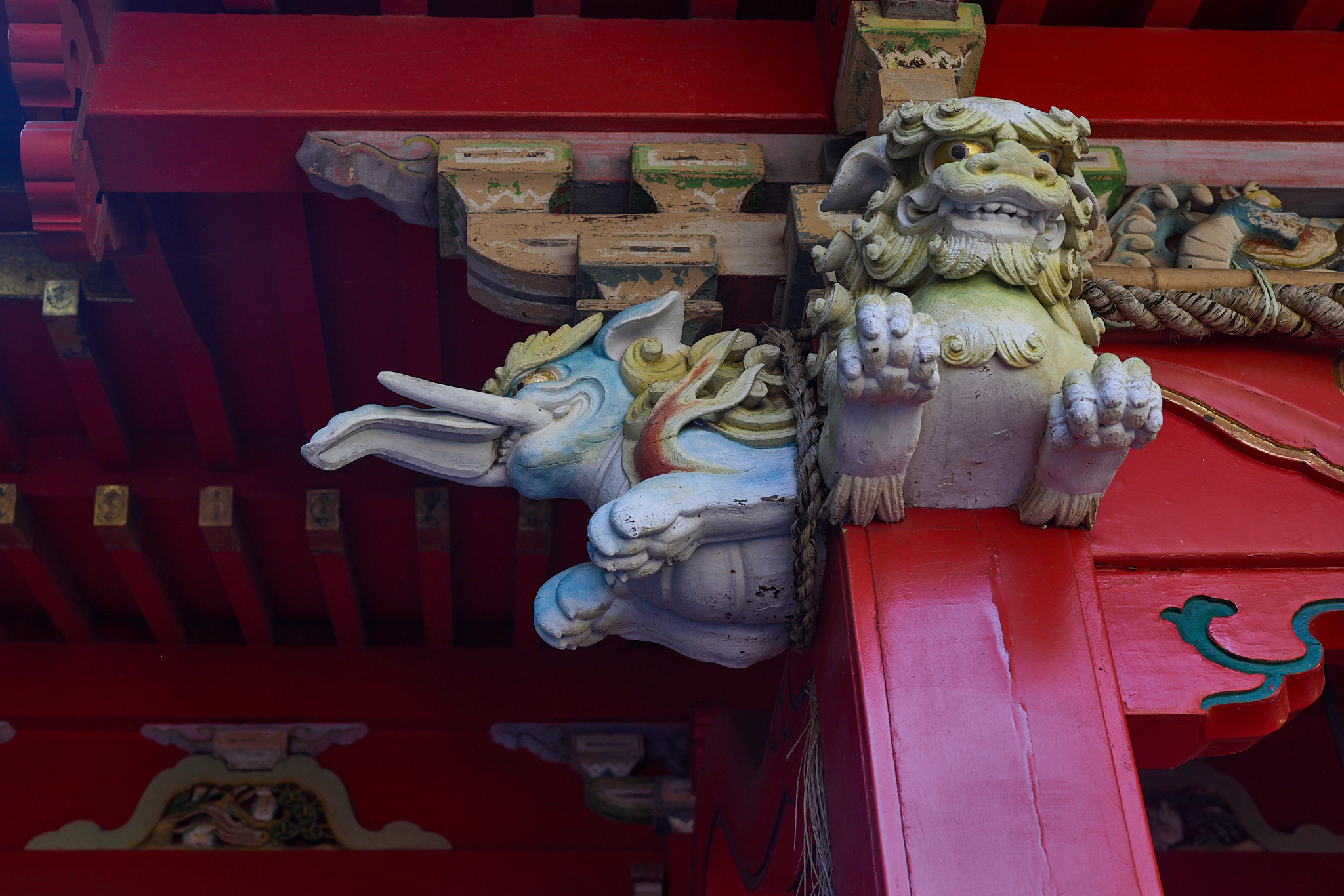 A carved lion supporting a beam beneath red rafters, beside a carved pale creature with a long curled trunk projecting from a red pillar